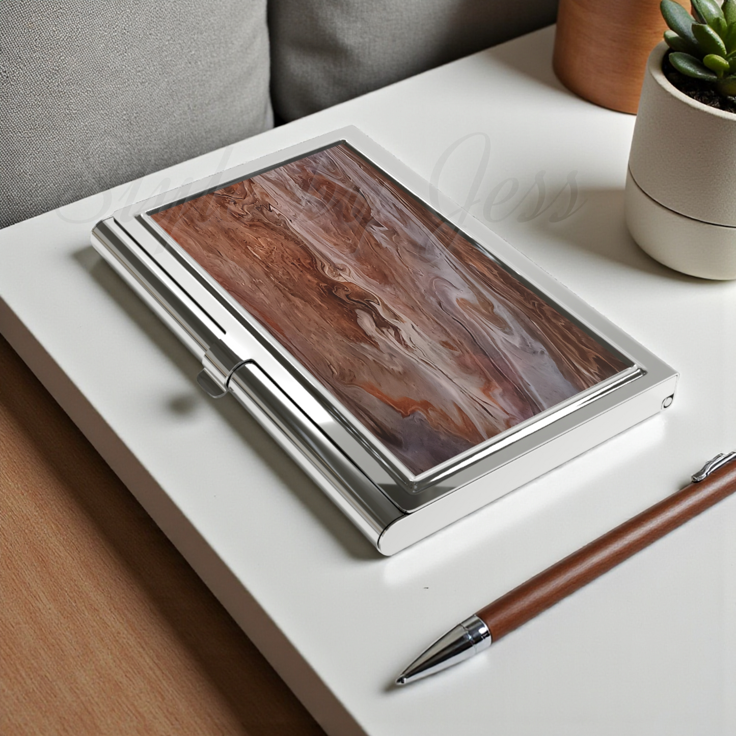 Wooden style business card holder with metallic cover on a white surface with a pen and plant in the background