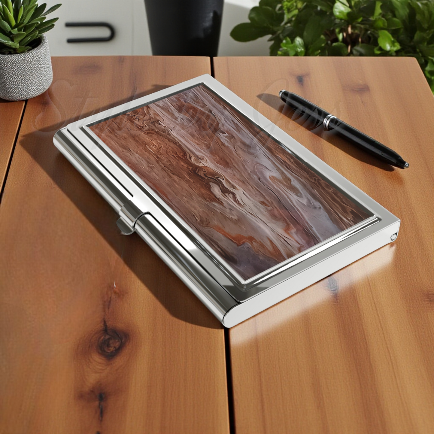Wooden desk with a art image mimicking wood on a silver business card holder designed for a professional gift, pen and potted plant.