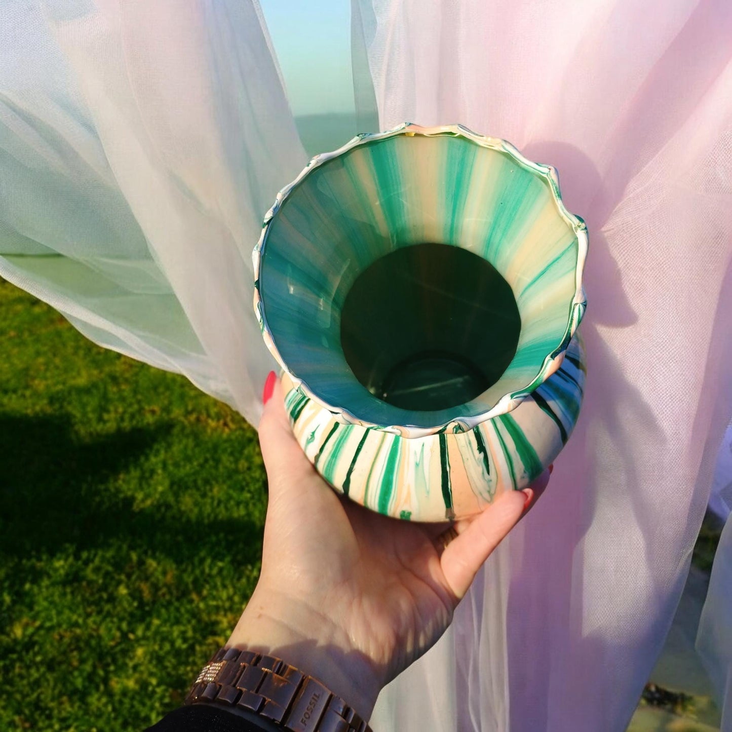 Hand holding a decorative vase with green and white stripes against a light fabric background.