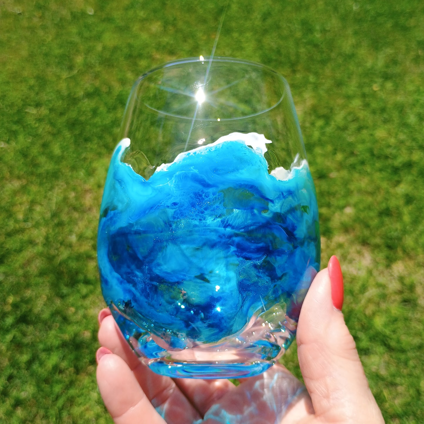 Hand holding a glass with blue and white design hand painted on the glass against a grassy background
