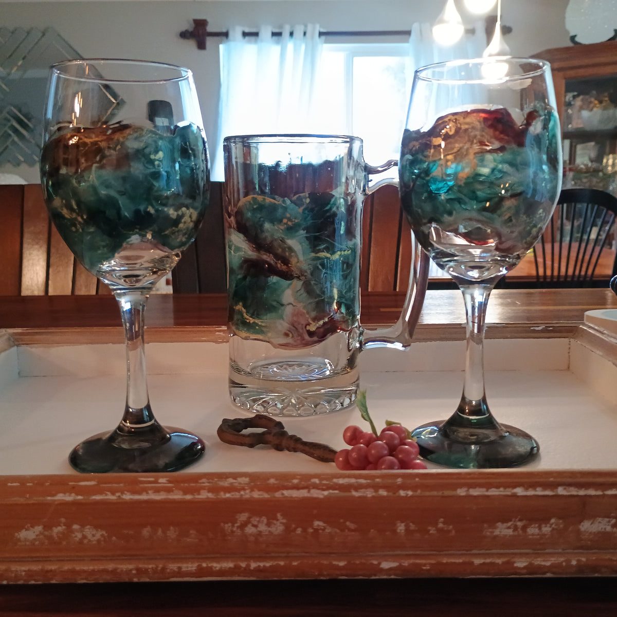 Two wine glasses and a mug showcasing the resin art design on each item sitting on a rustic wood tray with a decorative interior in the background. A style by Jess artistic creation for gift ideas.