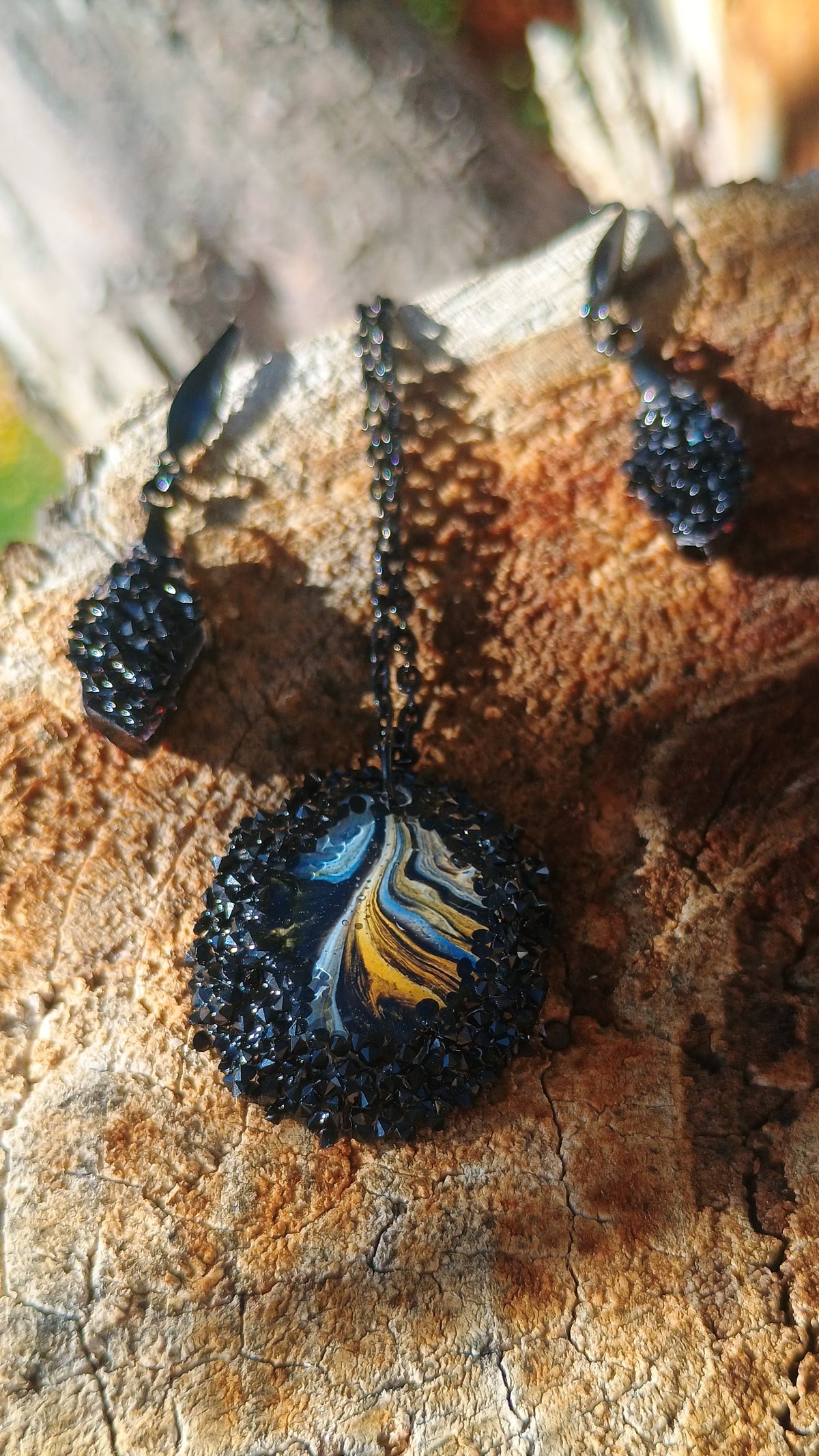 A black miniature painting pendant with black crystals surrounding the painting and covering the drop down dangle earrings. The pendant features a blue, white, and yellow feather design, displayed on a rustic wood log alongside matching drop earrings a Style By Jess at Gotta Have It Design 