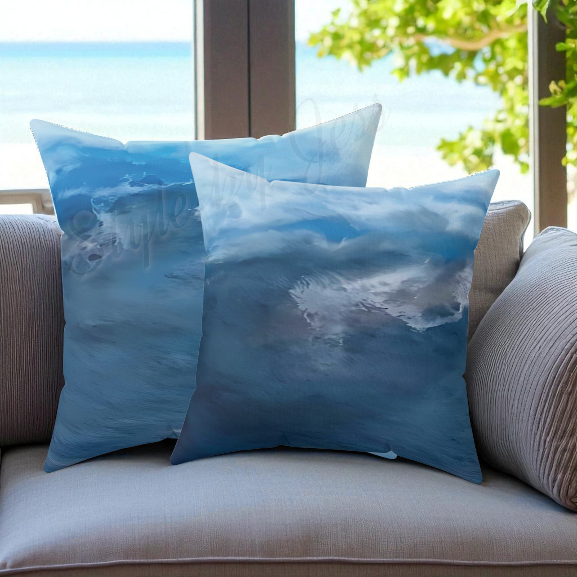 Two blue ocean-themed pillows on a couch with a window view of greenery and water.