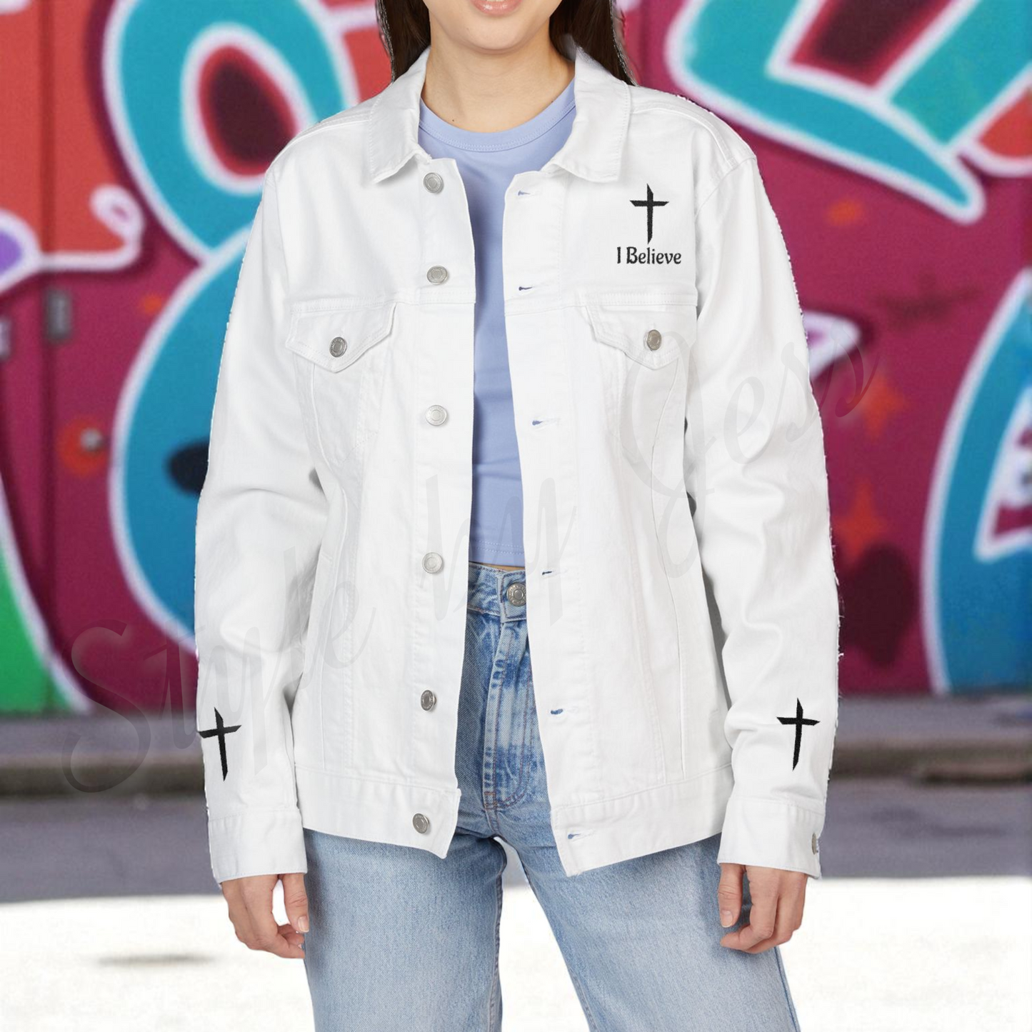 Person wearing a white denim jacket with crosses and 'I Believe' text in front of colorful graffiti.