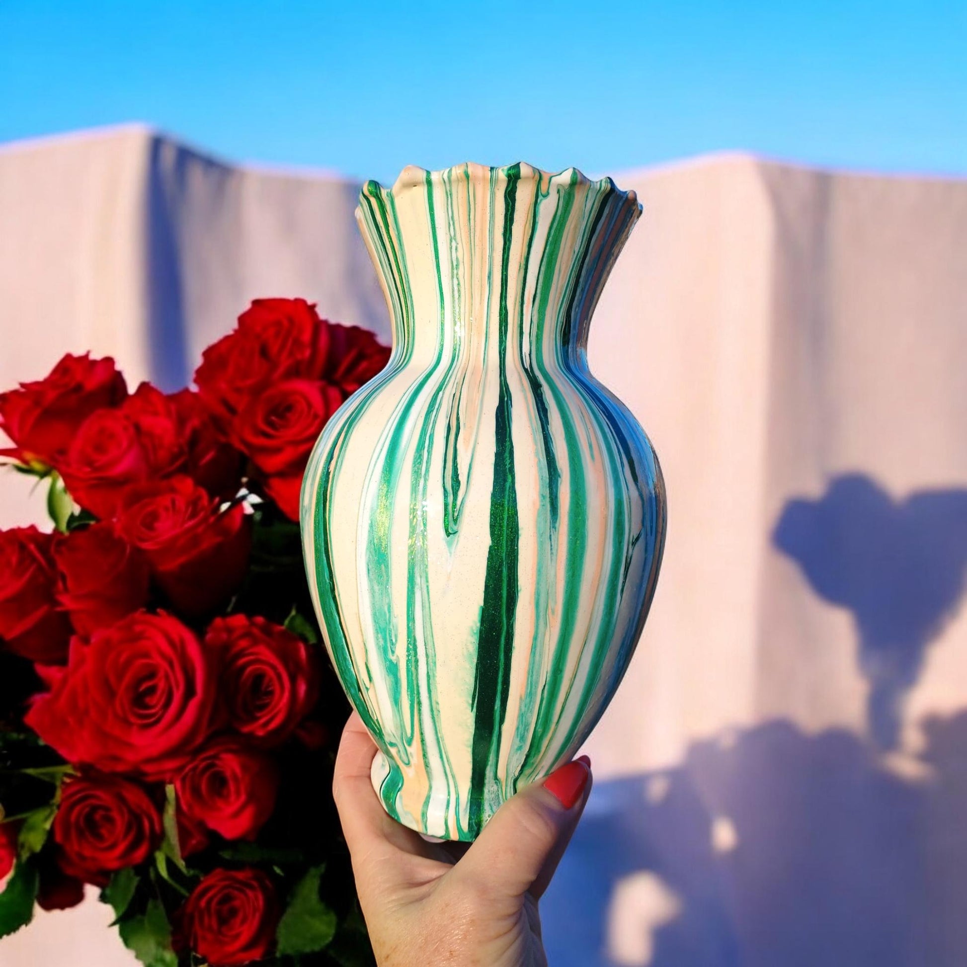 Hand holding a green and white striped vase with red roses against a blue sky.