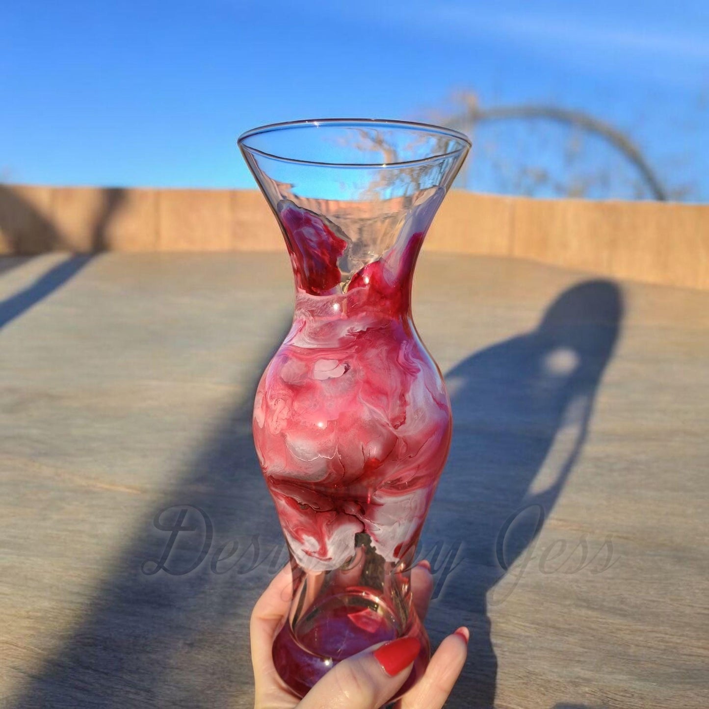 Glass vase with red and white marbled design held by a hand on a wooden surface with blue sky in the background.