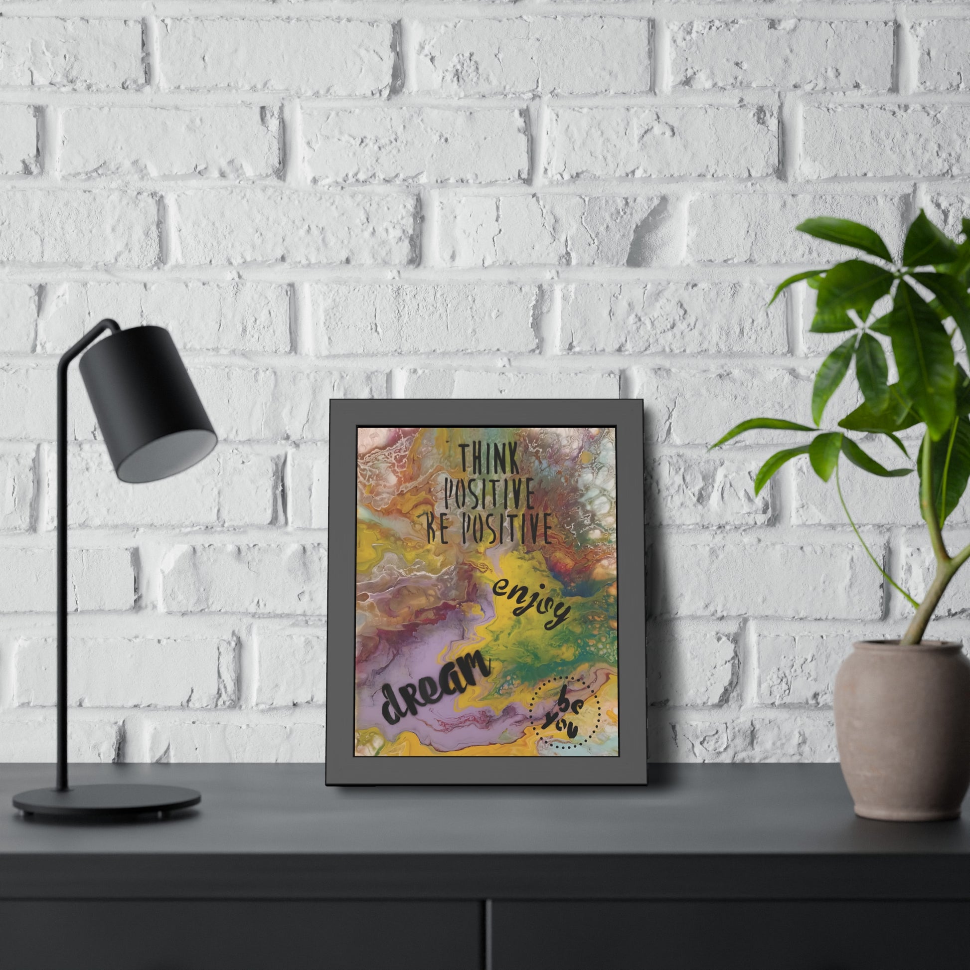 Framed motivational poster on a desk with a lamp and plant against a white brick wall.
