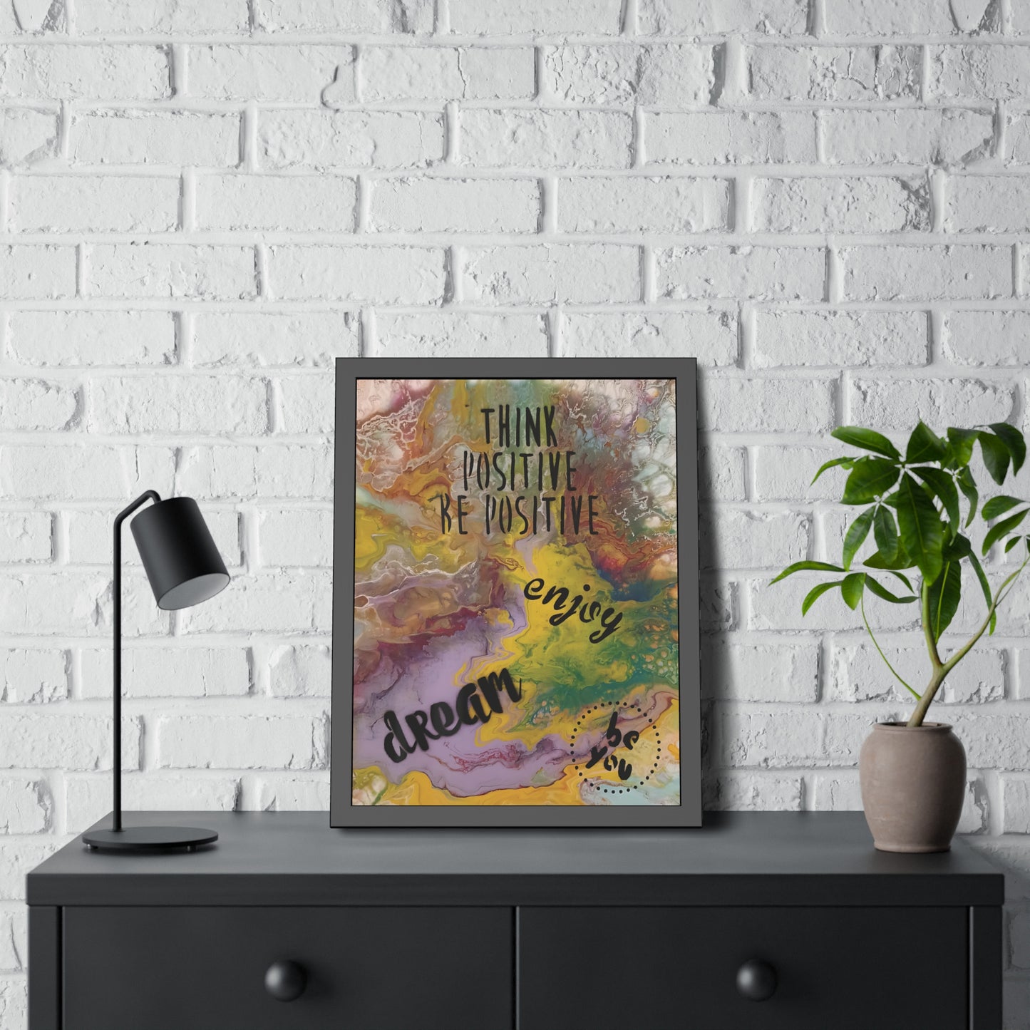 Framed motivational poster on a black dresser against a white brick wall with a lamp and plant.
