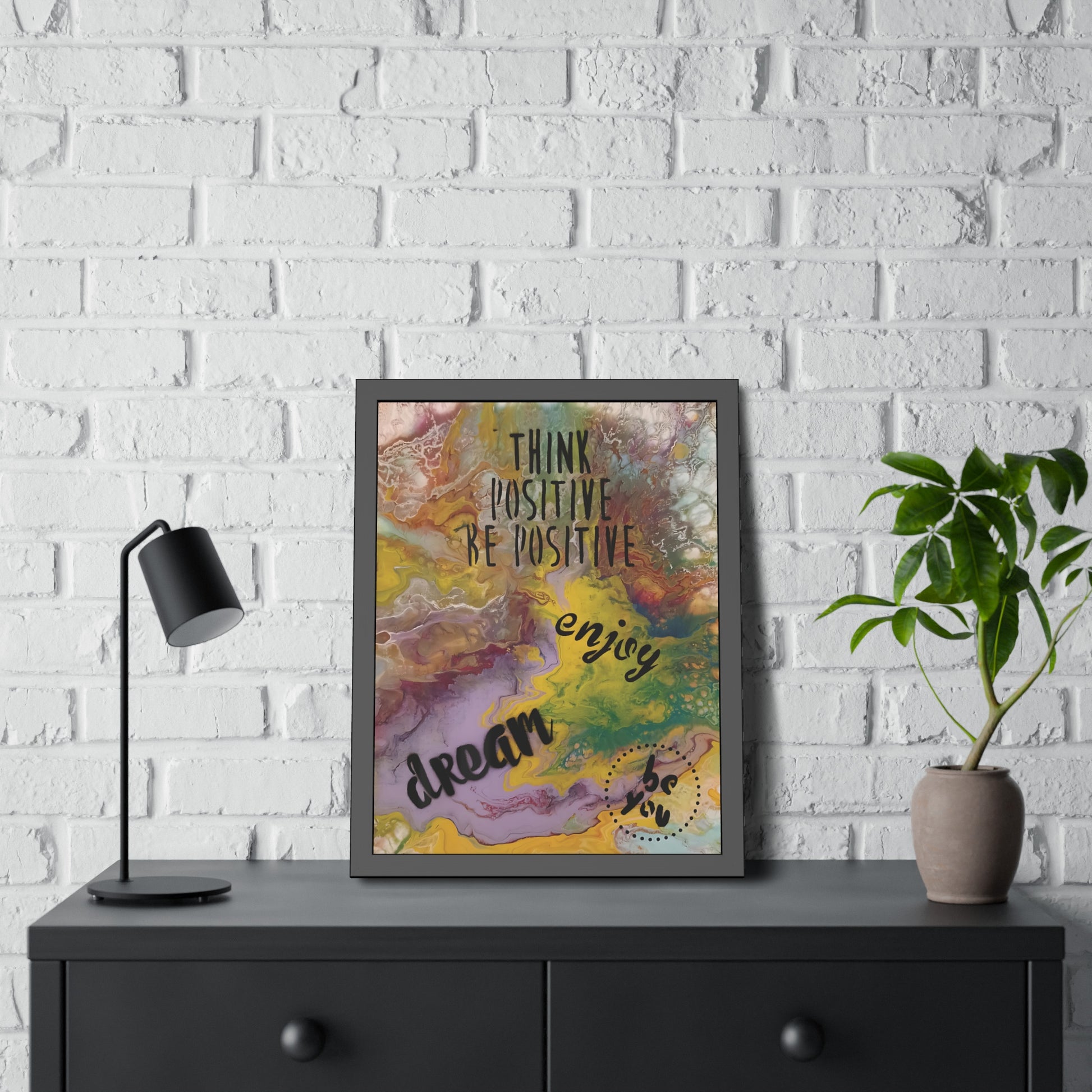 Framed motivational poster on a black dresser against a white brick wall with a lamp and plant.