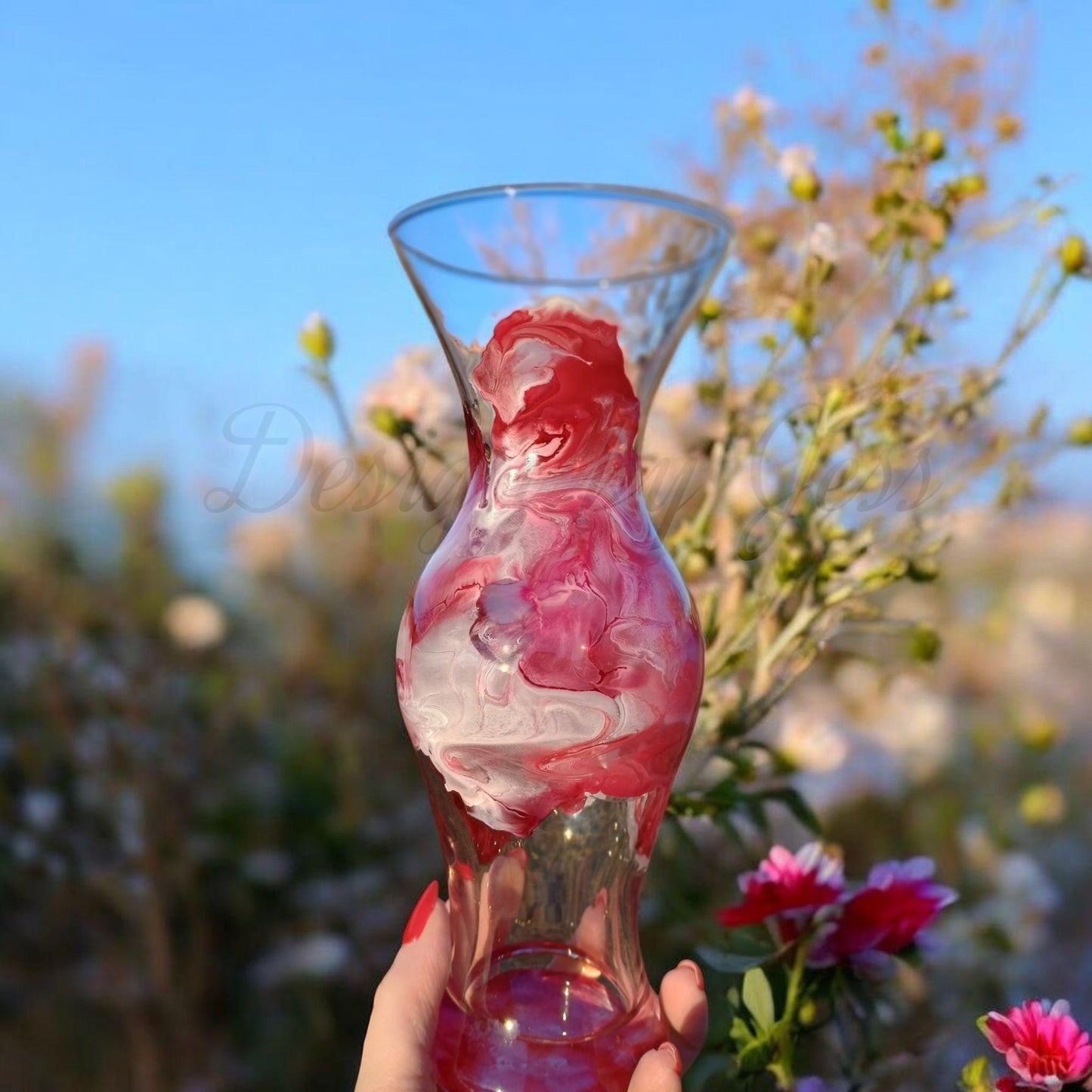 Glass vase with red and white design held in front of a natural background with flowers.