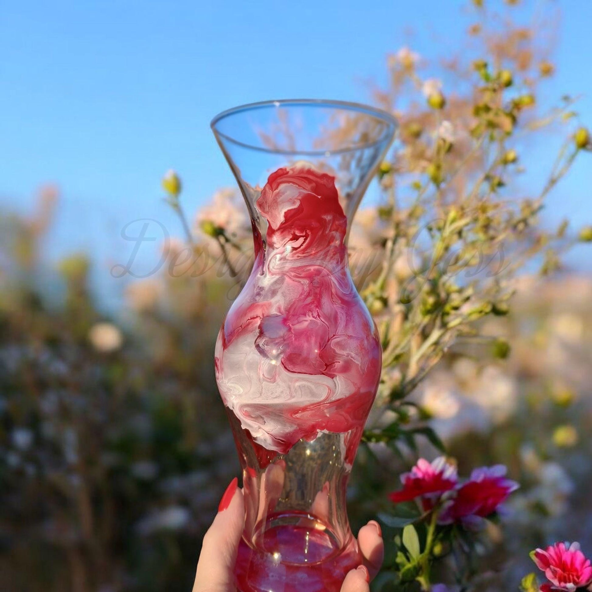 Glass vase with red and white design held in front of a natural background with flowers.