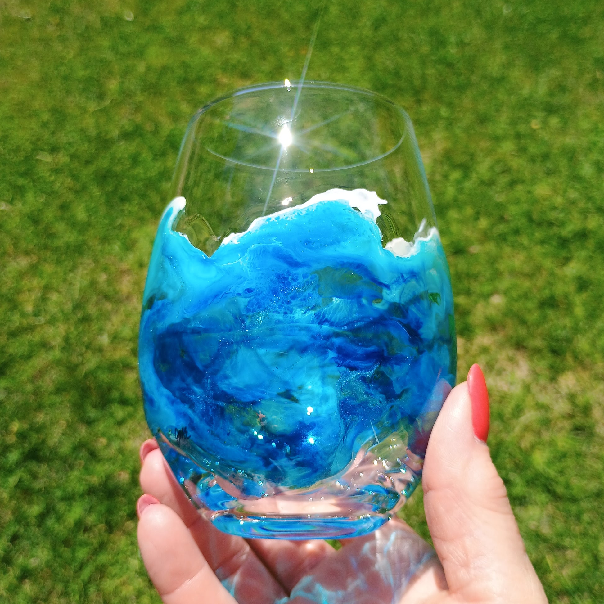 Hand holding a glass with blue and white design hand painted on the glass against a grassy background