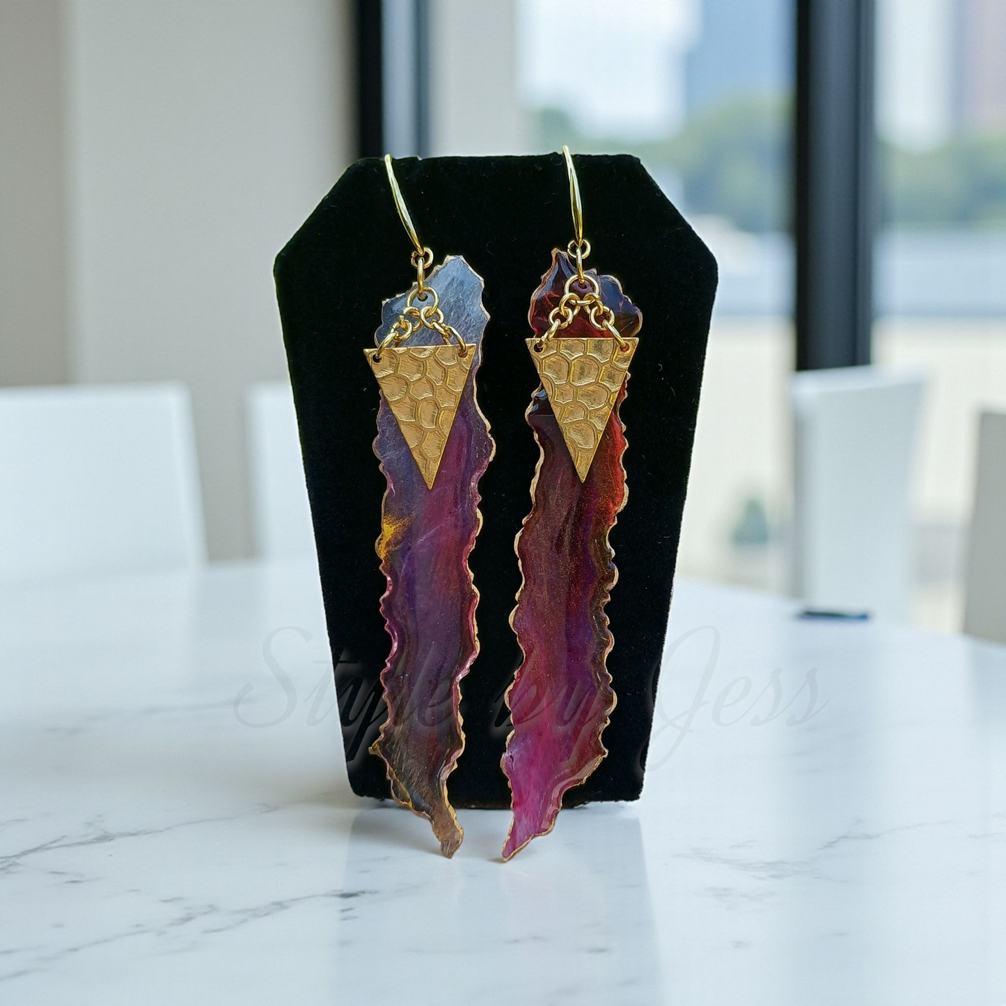 Pair of long, colorful earrings on a black stand with a blurred indoor background Style by Jess