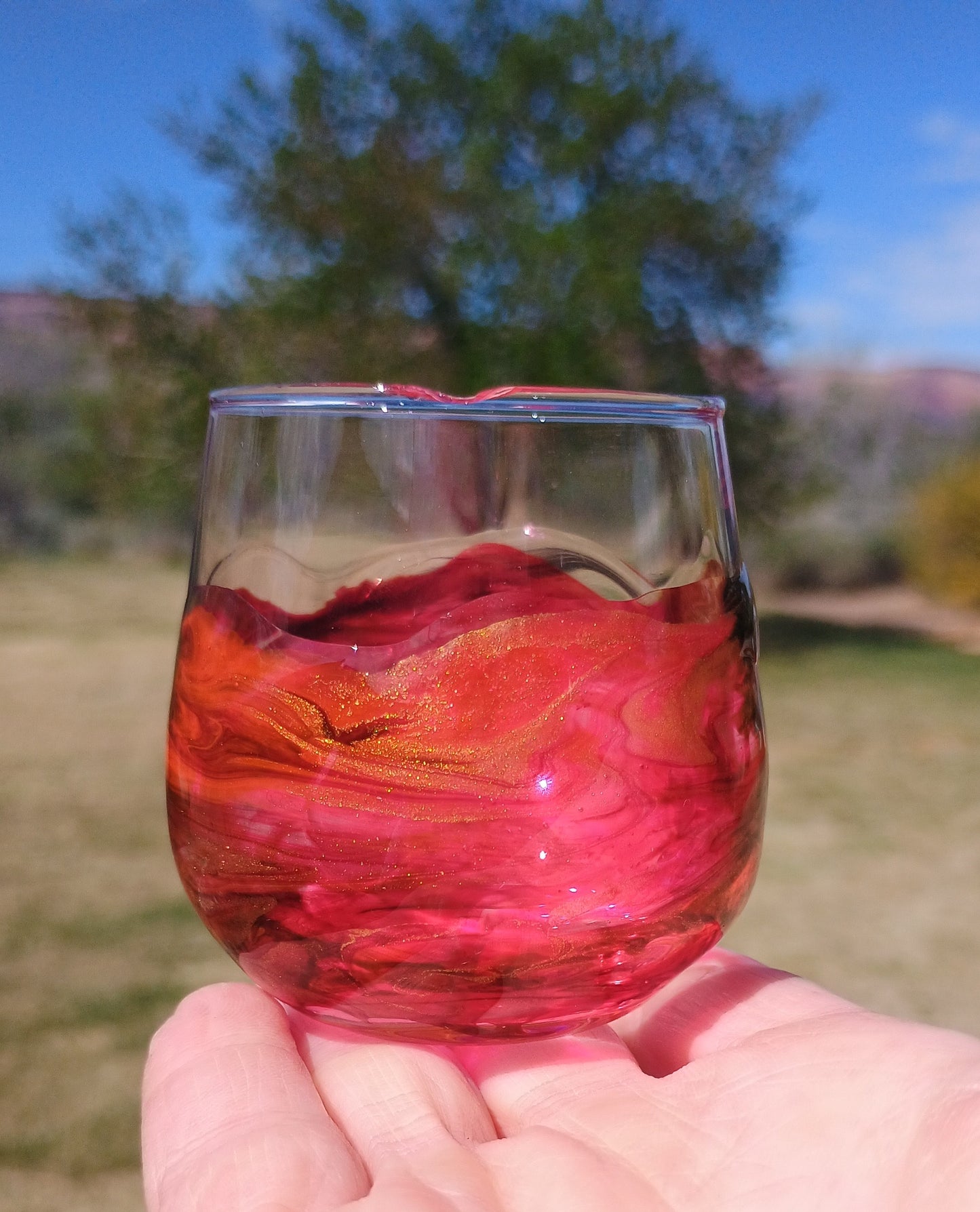 Hand holding a barware glass with red art against a blurred outdoor background