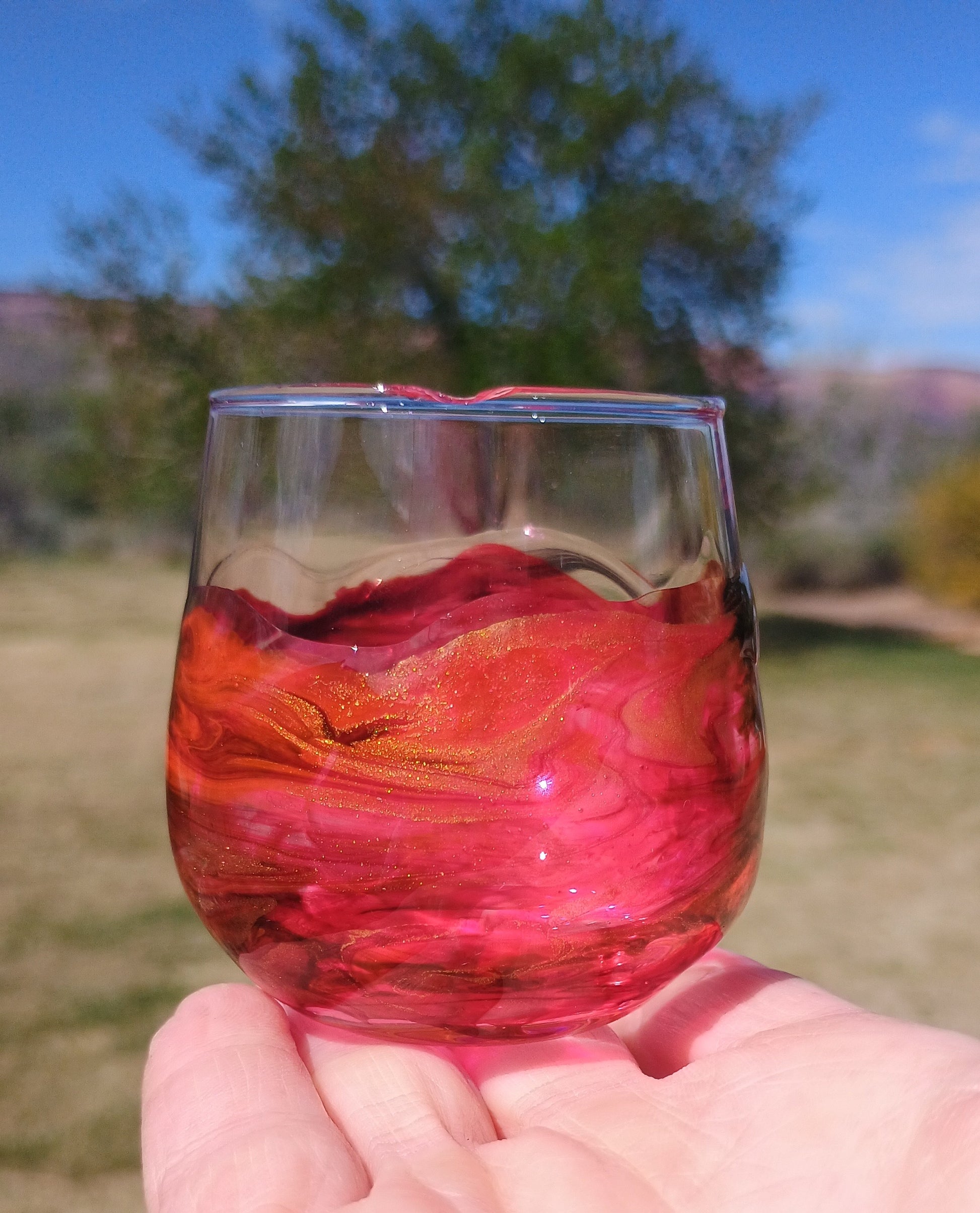 Hand holding a barware glass with red art against a blurred outdoor background