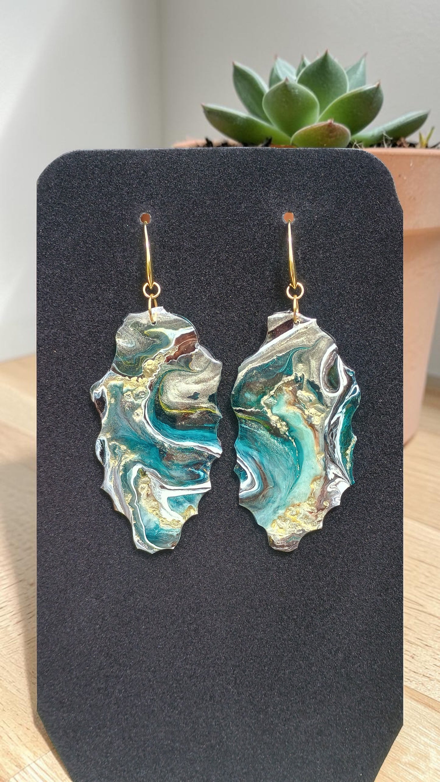 A set of earrings handmade teal, gold and white geode style resin art a Style by Jess at Gotta Have It Design on a black display with a succulent plant in the background. 