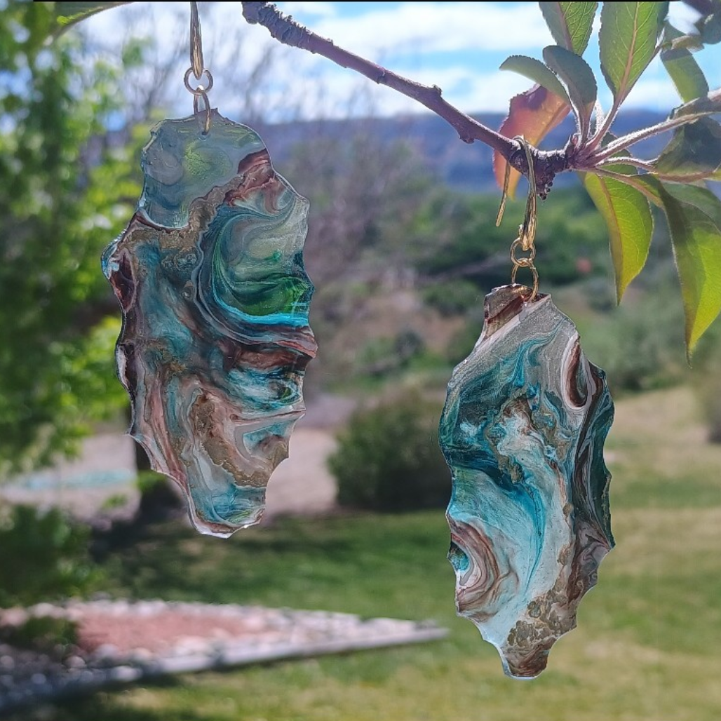 Pair of earrings with a geometric handmade design with swirls of blue teal, browns, gold and white and slightly transparent a custom set created by Style By Jess at Gotta Have It Design. The background is of green grass, green tree and mountains. 