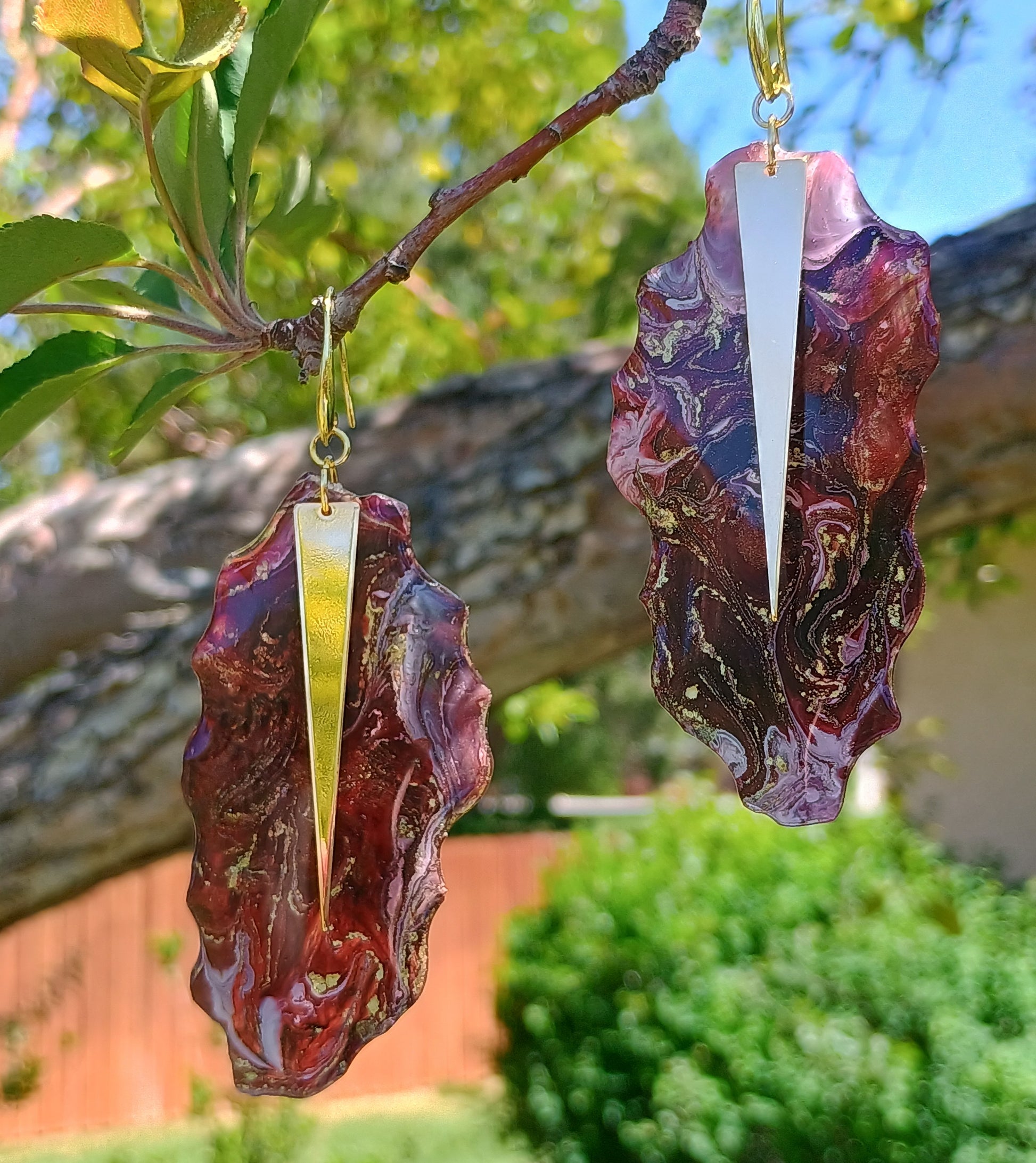 Red hand poured resin and ink  artistic earrings with 18 K Gold Plated gold hooks displayed on a branch outdoors. Style By Jess
