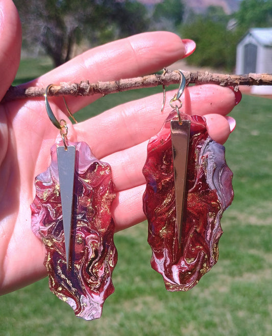 Red and gold marbled earrings held by a hand with a blurred green outdoor background a custom fashion piece a Style By Jess at Gotta Have It Design. 