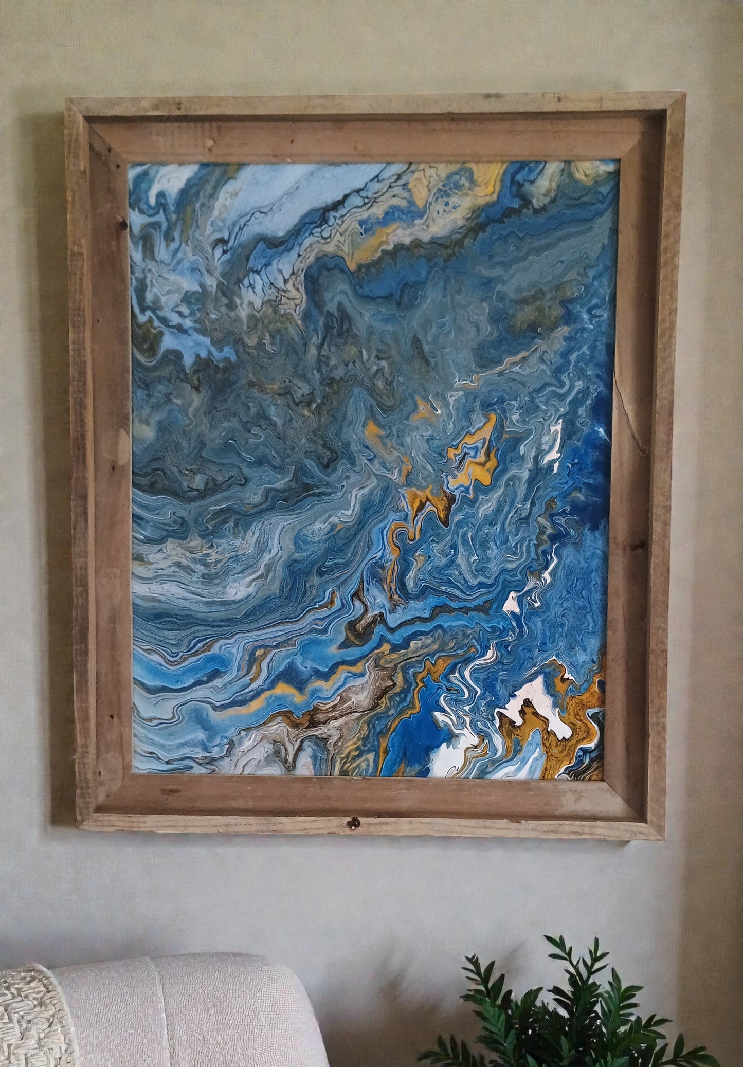 Framed abstract artwork with blue and gold swirls hanging on a wall in a minimalist room with a white chair and a plant. 