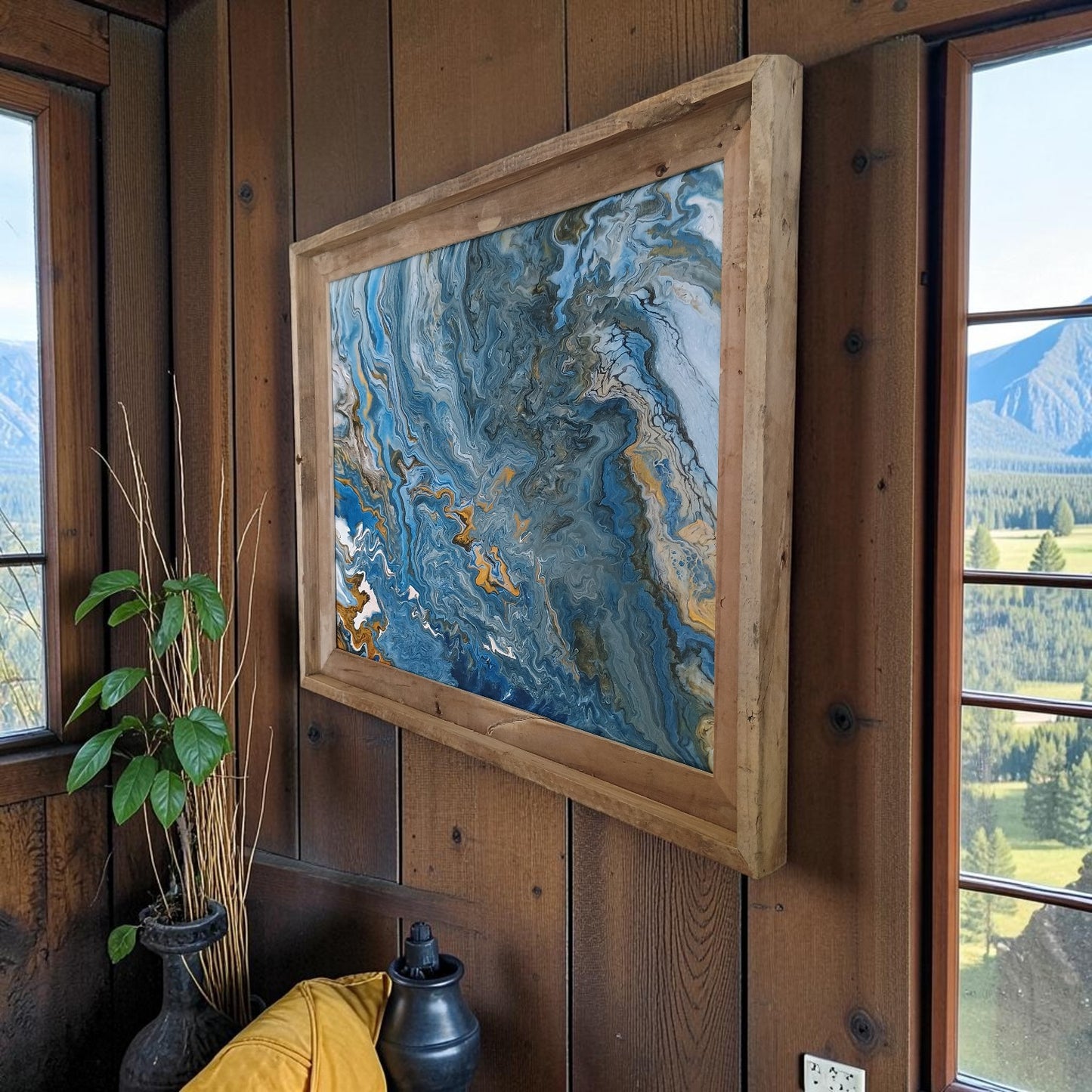 Abstract painting in a wooden frame on a wooden wall with windows showing a scenic view.