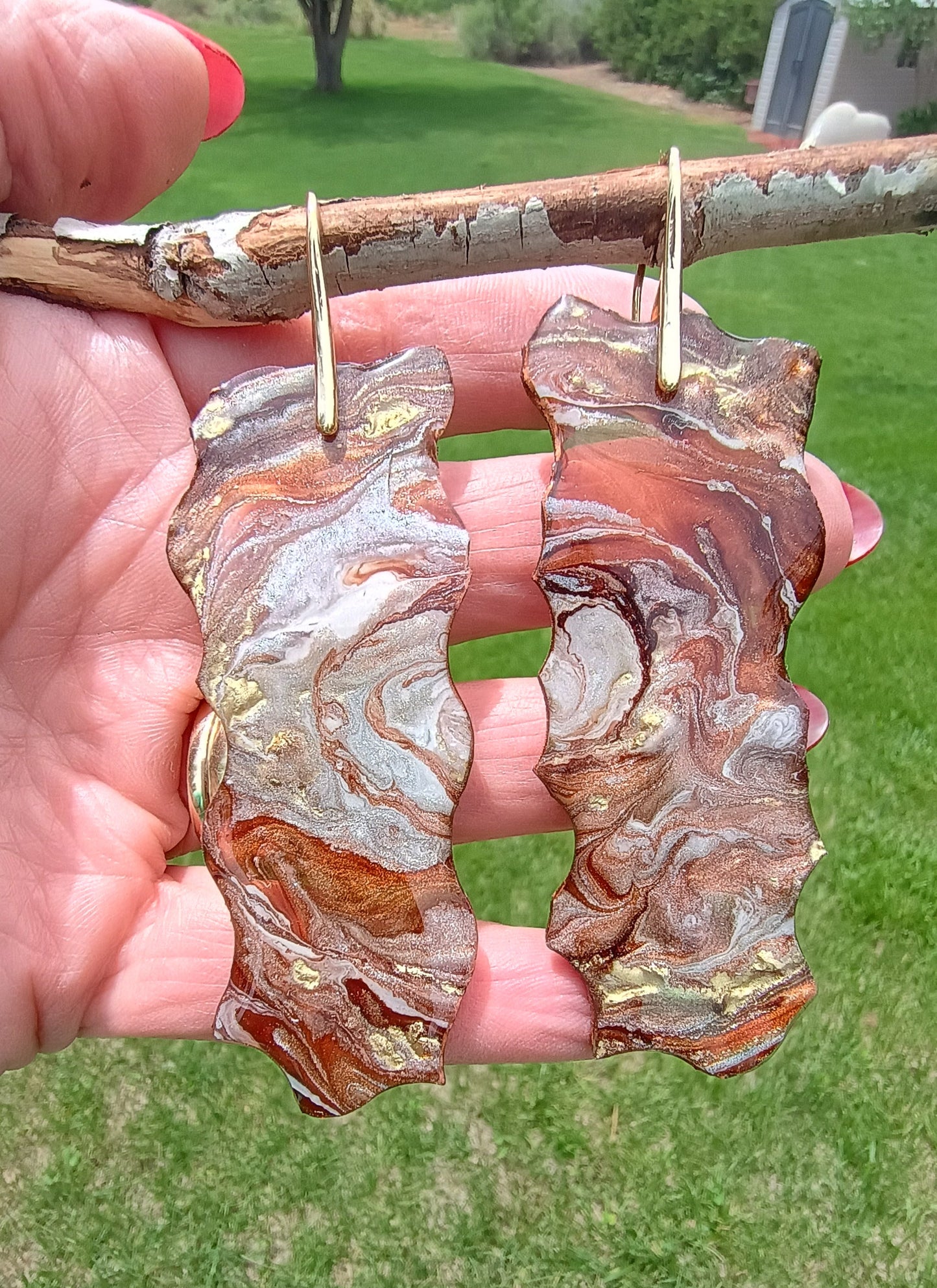 Decorative dangle earrings with earth tones and gold flowing with a marble like effect held in hand with grassy background