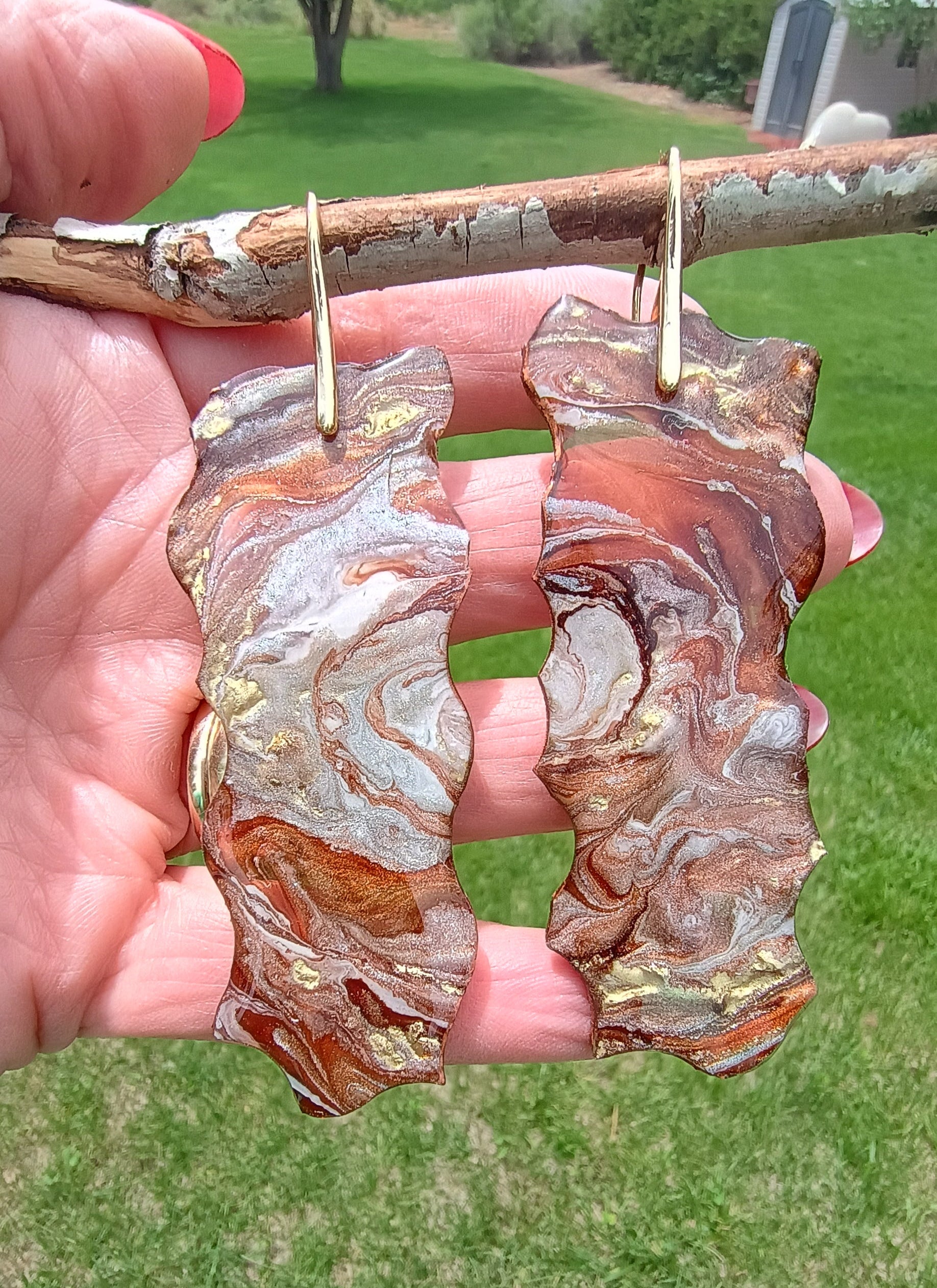 Decorative dangle earrings with earth tones and gold flowing with a marble like effect held in hand with grassy background