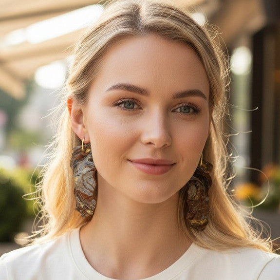 Woman wearing gold and brown stone looking resin dangle earrings sitting outdoors at a cafe with a blurred background