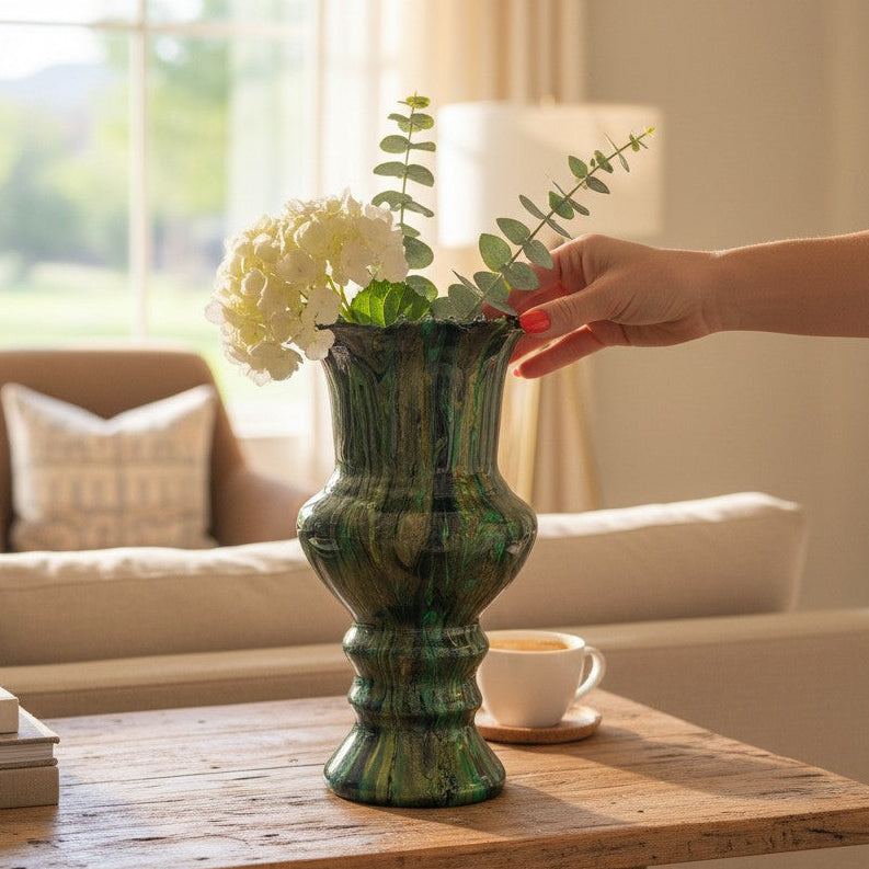 Person arranging flowers in an abstract artistic green and gold vase on a wooden table in a living room.
