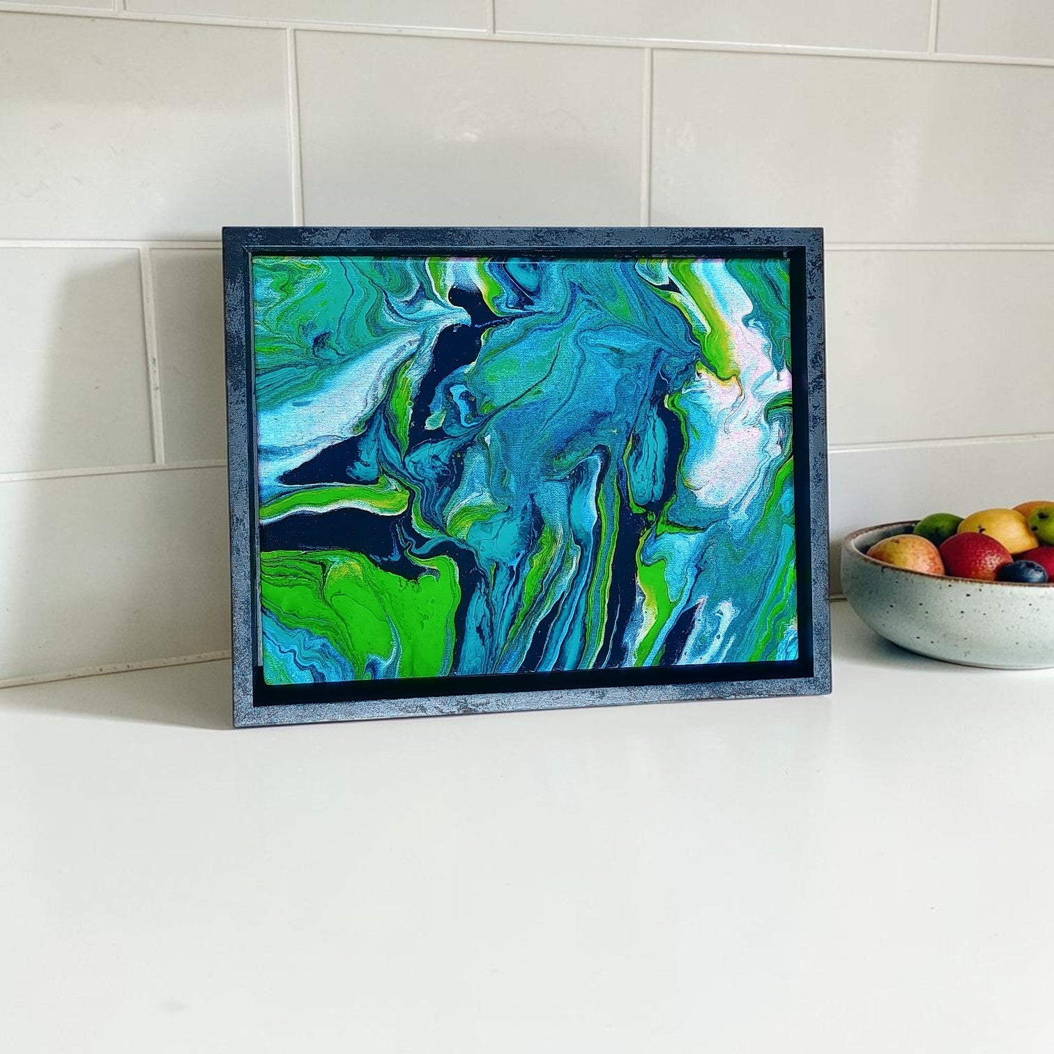 Abstract art piece in a frame on a white surface with a bowl of fruit in the background on display as kitchen decor. 