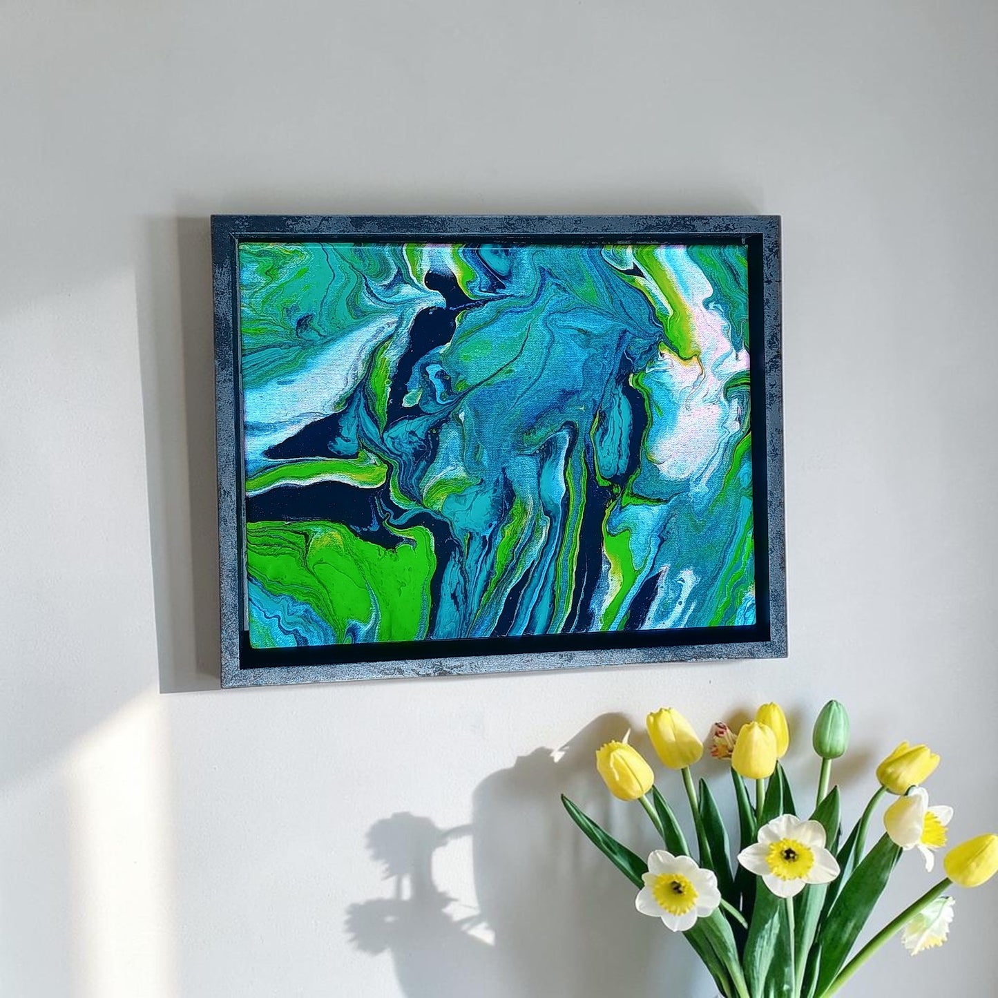 Framed abstract painting with swirling green, blue, and white colors on a white wall, accompanied by a bouquet of yellow and white flowers in a entry way.