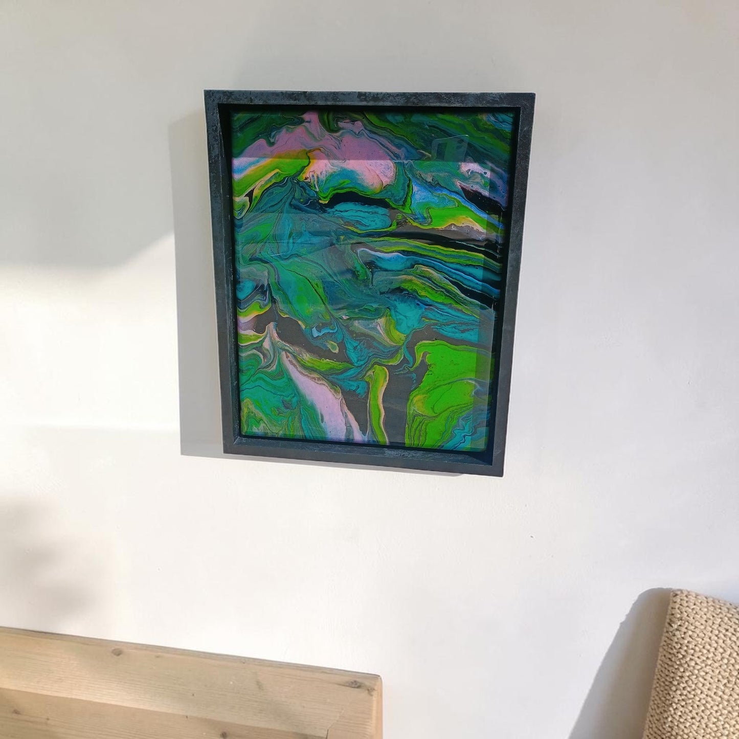 Framed abstract painting with green and blue colors on a white wall above a desk in a office.