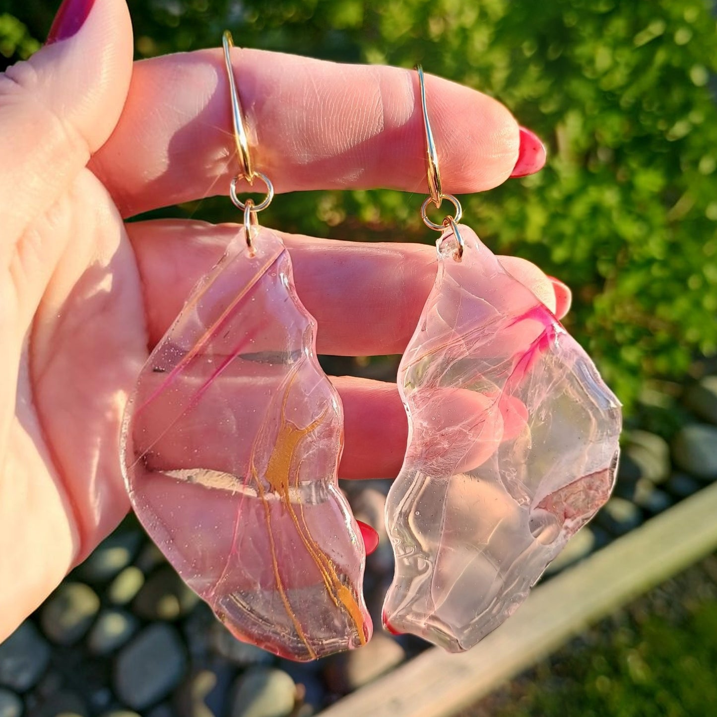 Pink and gold earrings held in a hand with a blurred green background