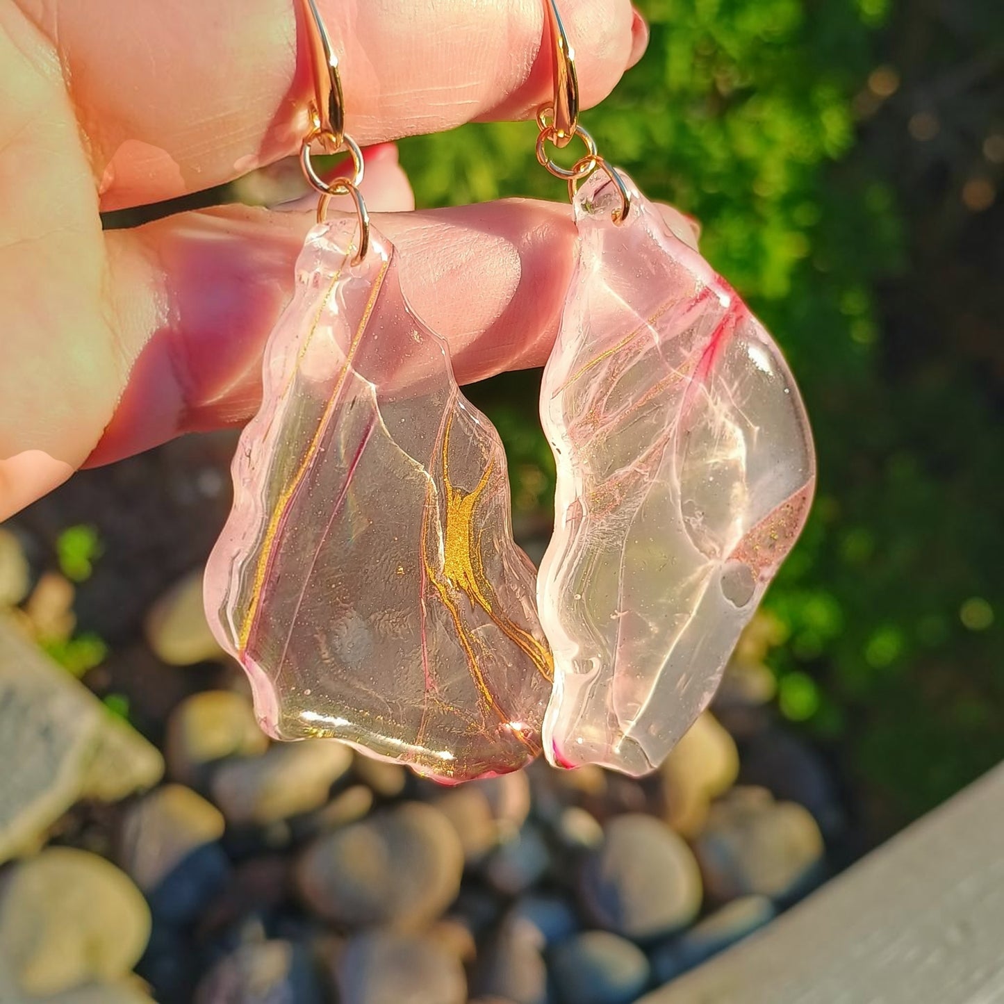 Handmade transparent resin dangle earrings held in natural golden hour sunlight, showing luminous clarity with gold streak, blush pink, and earth tone swirls — 18K gold plated hooks — one of a kind wearable art by Style By Jess