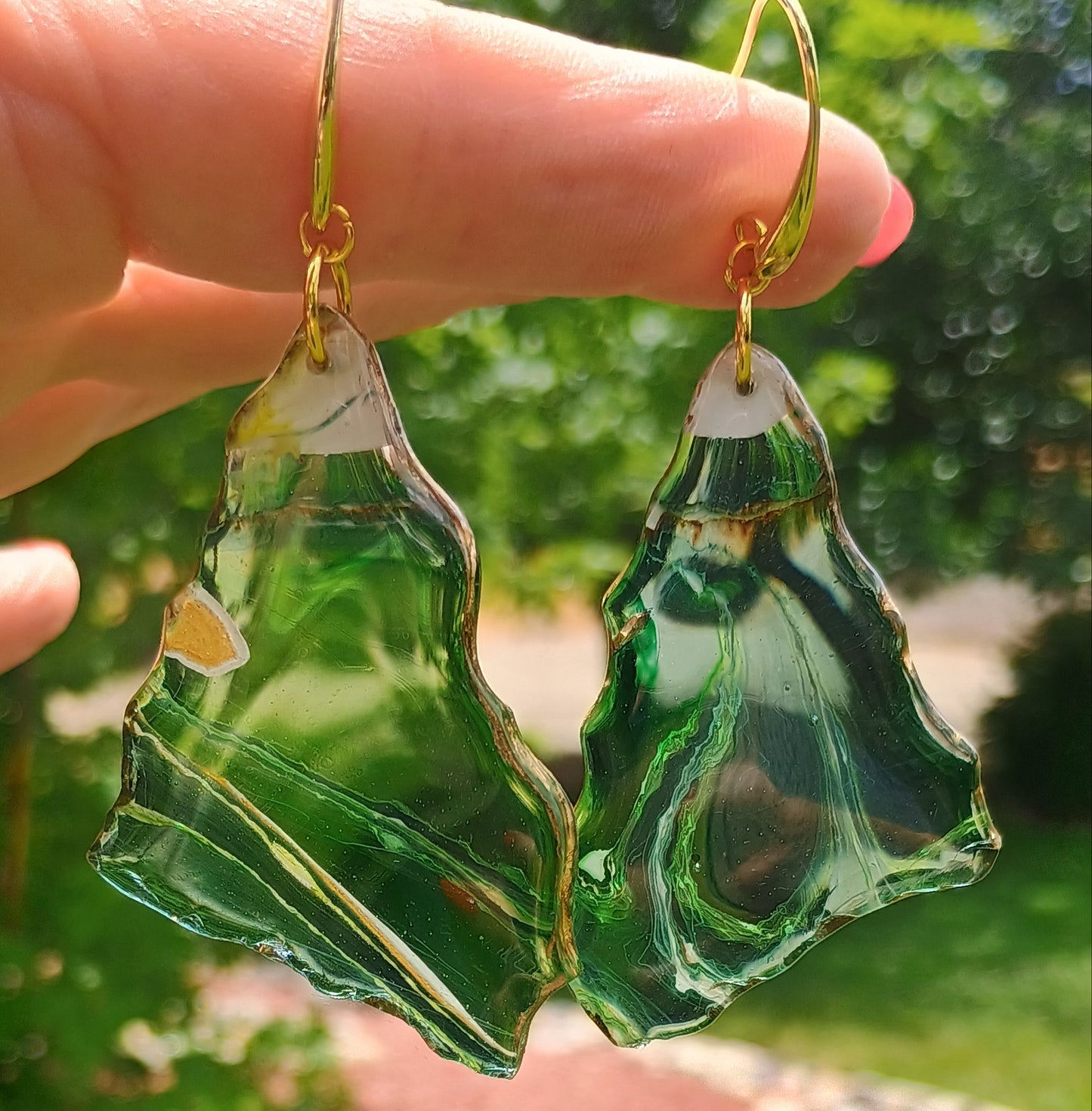 Green leaf-shaped earrings held by a hand with a blurred green outdoor background