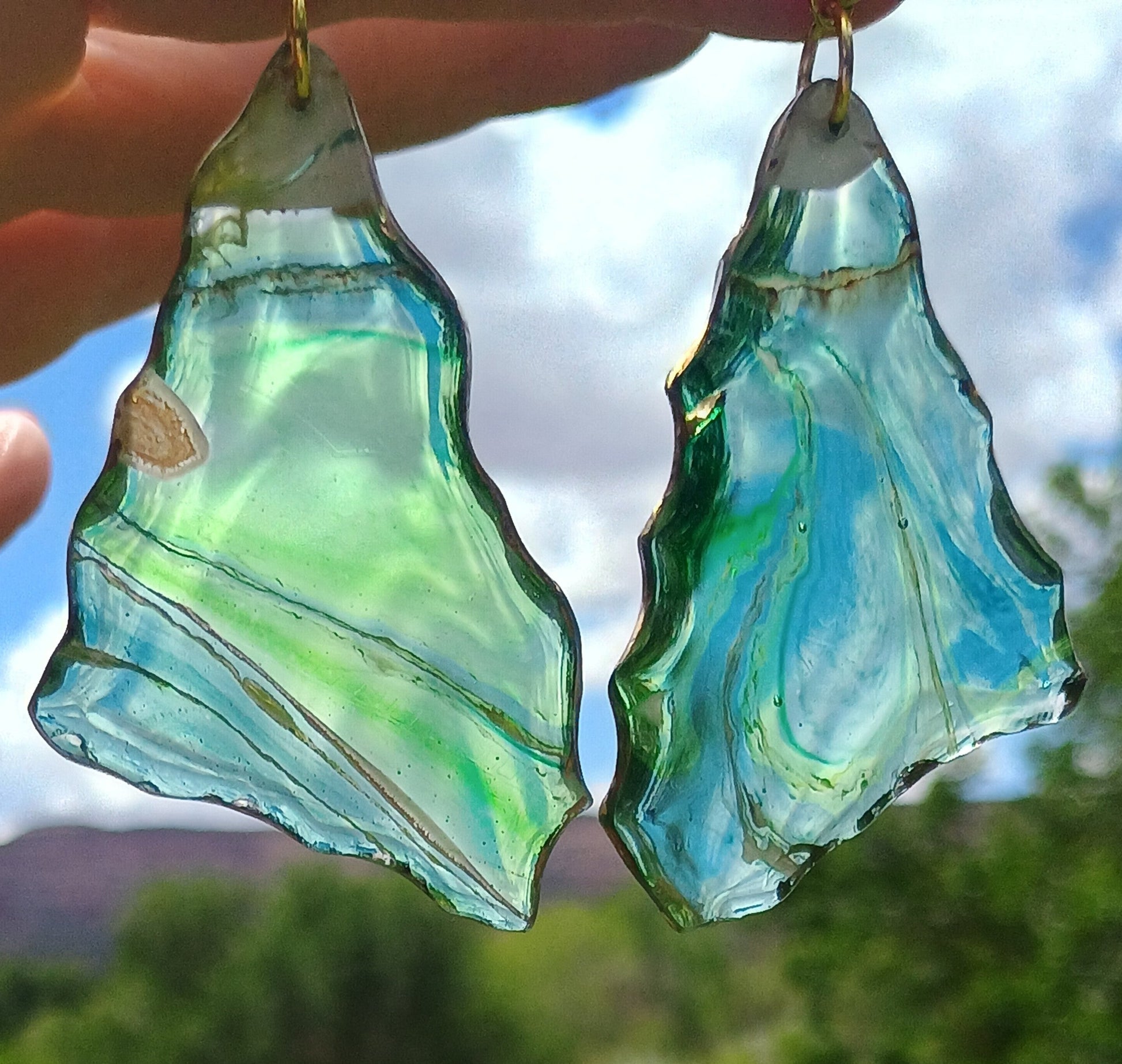 Handmade resin art dangle earrings held up to natural light — translucent evergreen base with gold, yellow, white and brown ribbon swirls and snow cap mountain design, featuring 18K gold plated ear wires, by Style By Jess at Gotta Have It Design