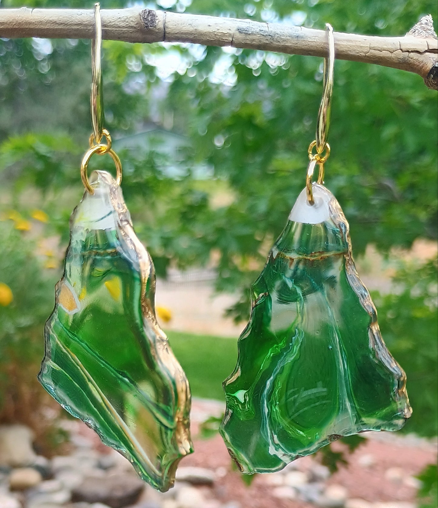 Dangle earrings with a green transparent base and veins of gold, brown and white throughout and a gold edge trim hanging from a branch with a natural background.  A Style By Jess at Gotta Have It Design. www.StyleByJess.com