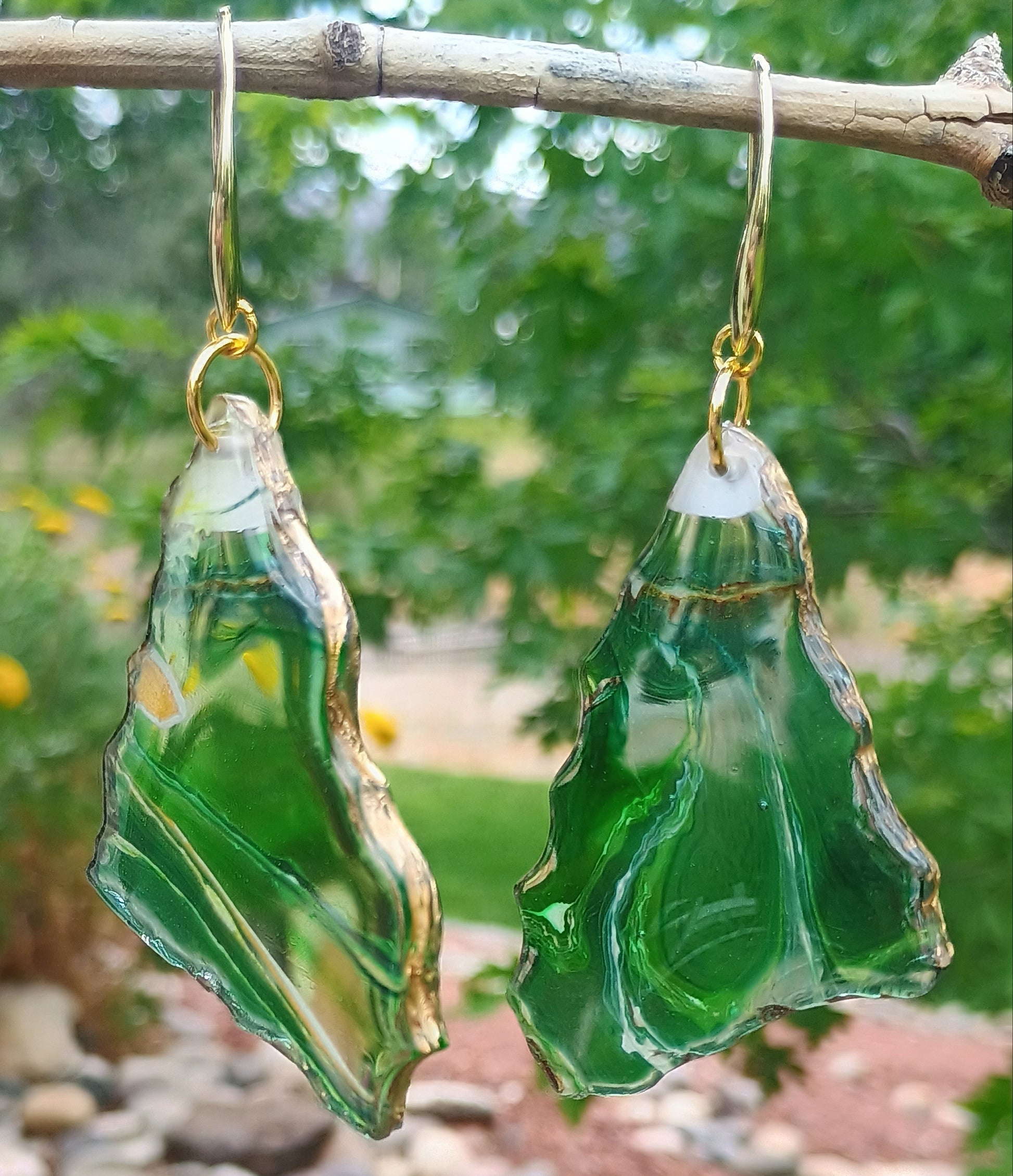Dangle earrings with a green transparent base and veins of gold, brown and white throughout and a gold edge trim hanging from a branch with a natural background.  A Style By Jess at Gotta Have It Design. www.StyleByJess.com