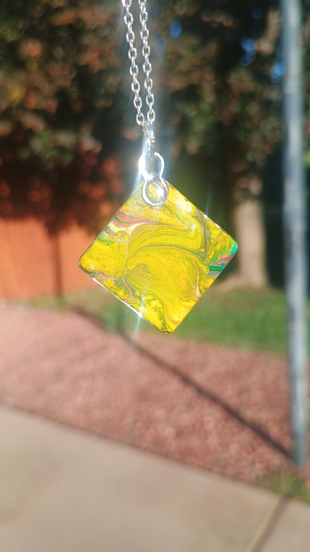 A square pendant necklace with a marbled yellow, green, and white pattern, suspended from a silver chain against a blurred outdoor background.