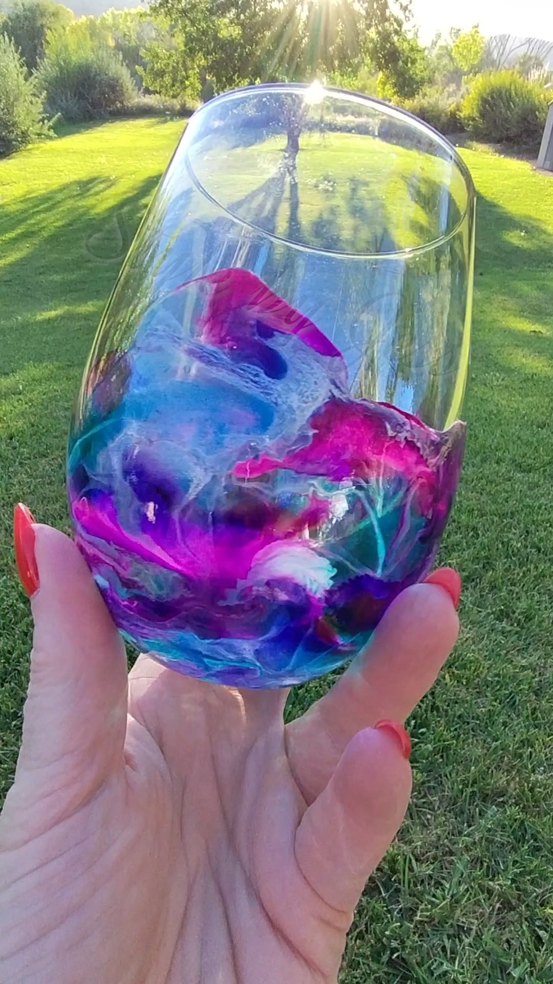 Hand holding a colorful glass with abstract design against a grassy background Style by Jess