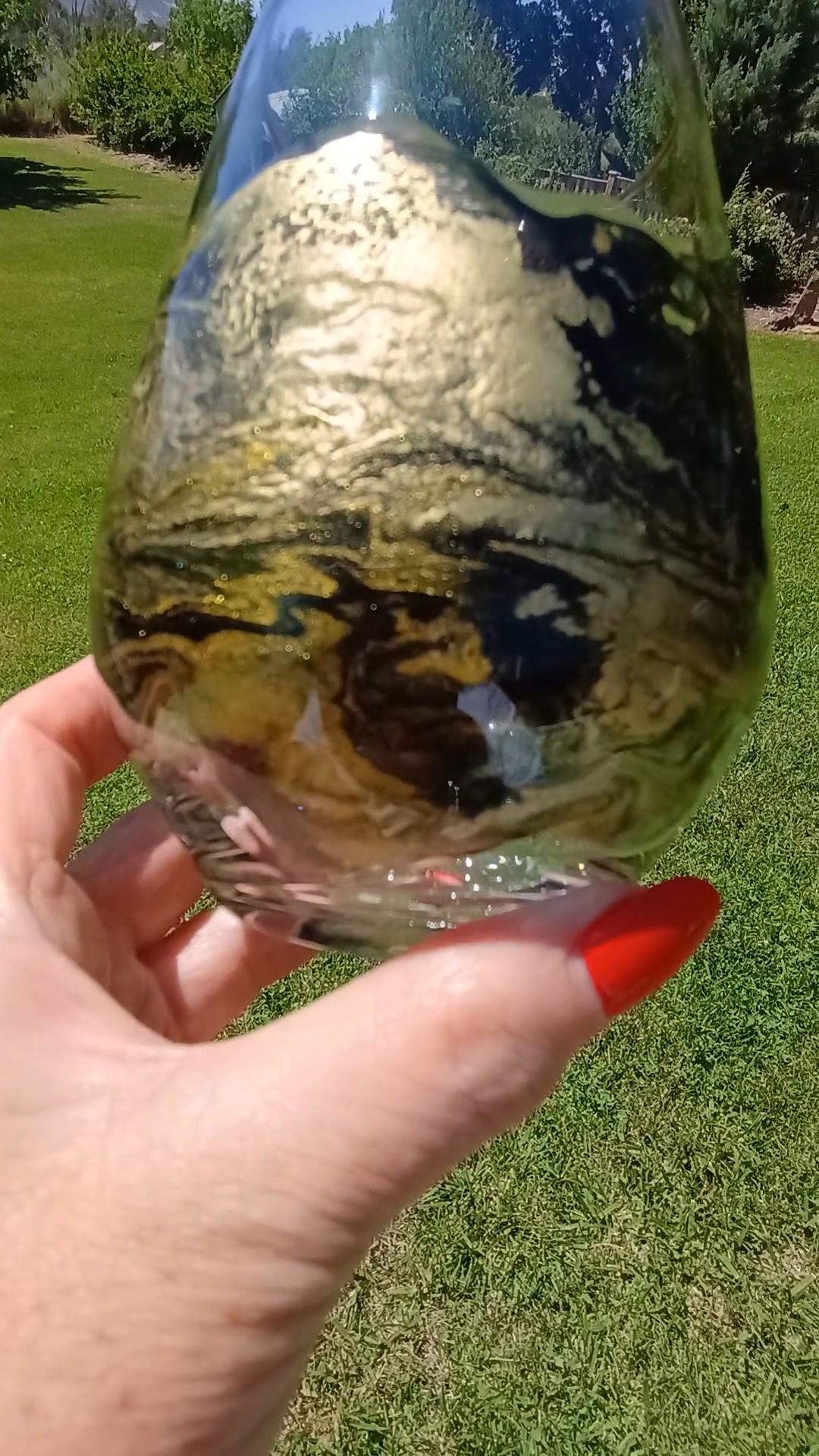 Hand holding a glass with black and gold abstract resin art on it against a grassy background