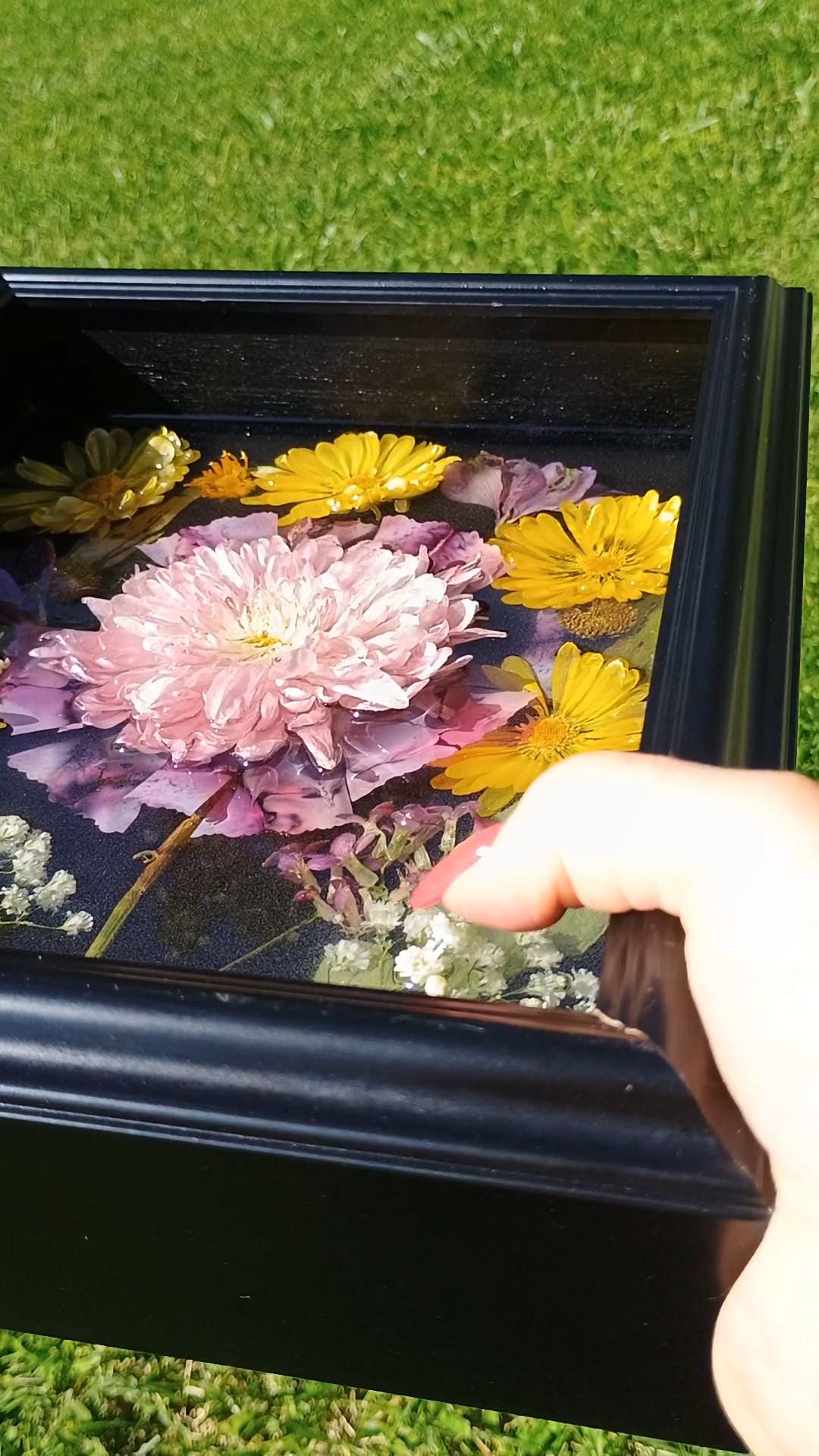 Hand holding a black frame with dried flowers in resin against a grass background