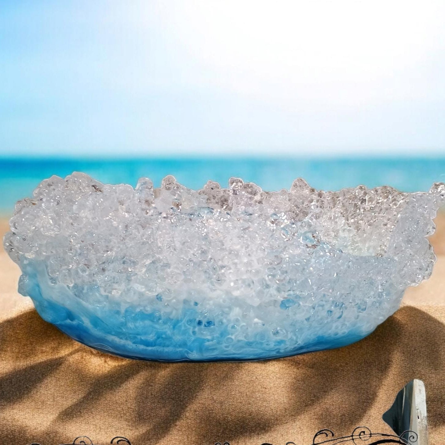 Blue decorative bowl on sand with ocean background, 'Design by Jess' text overlay
