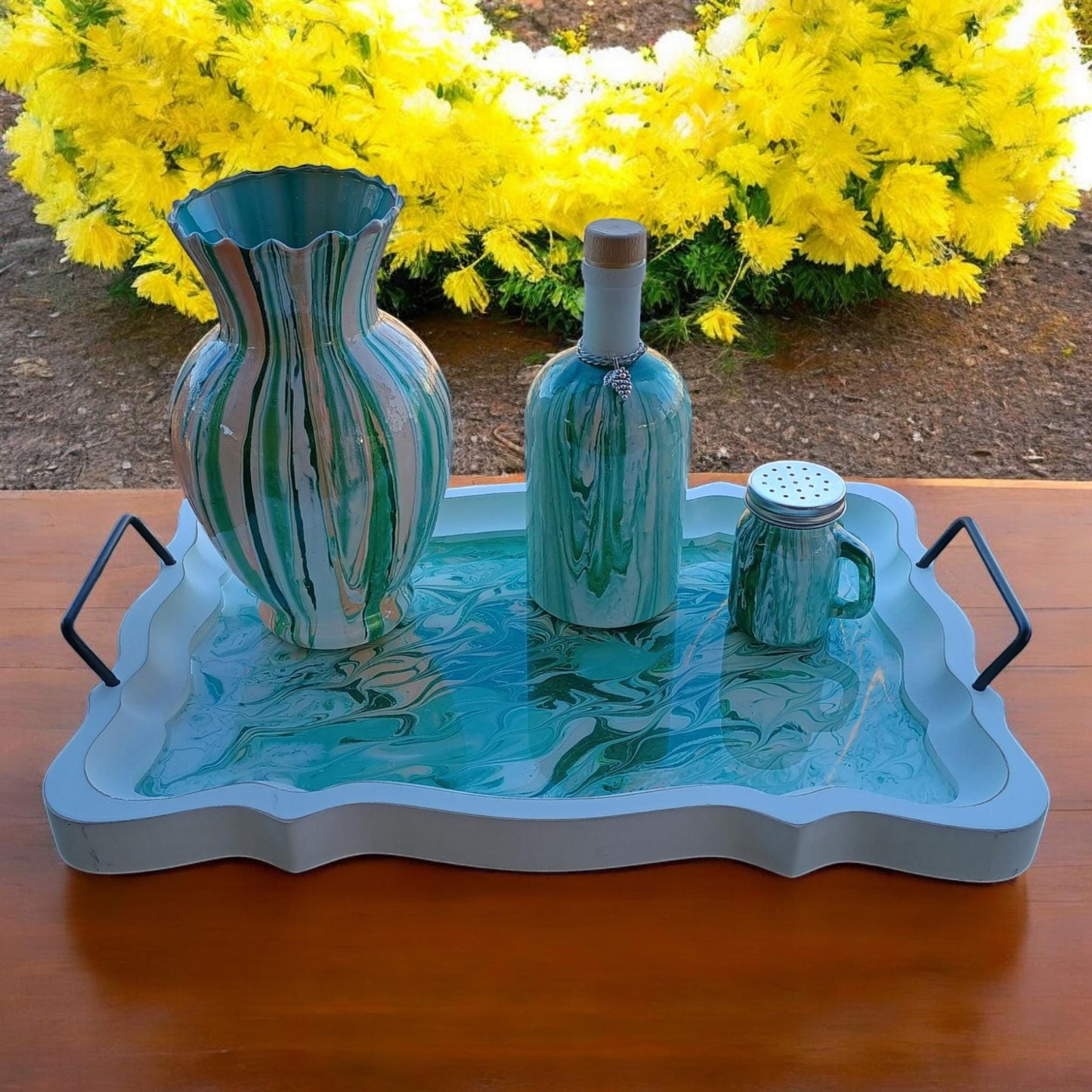 Decorative tray with a vase, bottle, and salt shaker on a wooden surface with yellow flowers in the background.