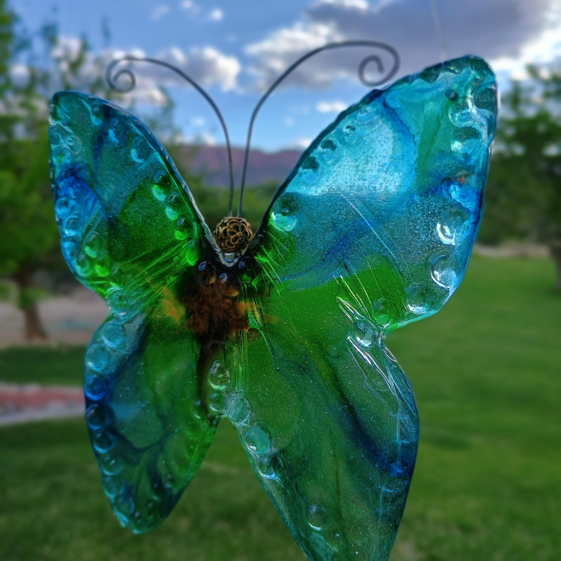 Glass butterfly sun catcher with blue and green wings on a blurred outdoor background a handmade creation by Jess with Style By Jess at Gotta Have It Design