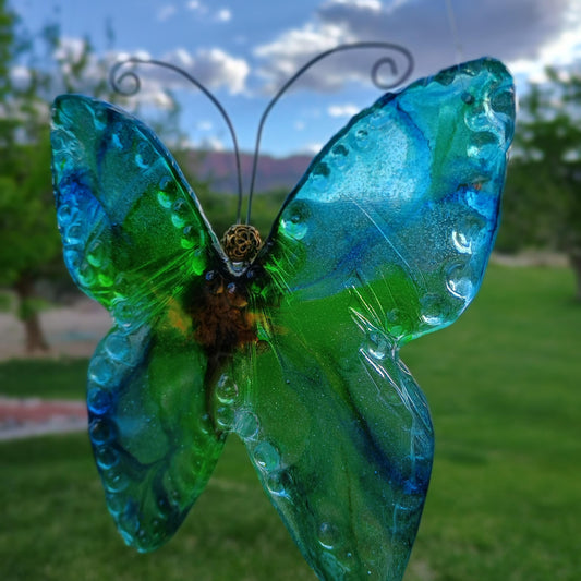 Glass butterfly sun catcher with blue and green wings on a blurred outdoor background a handmade creation by Jess with Style By Jess at Gotta Have It Design