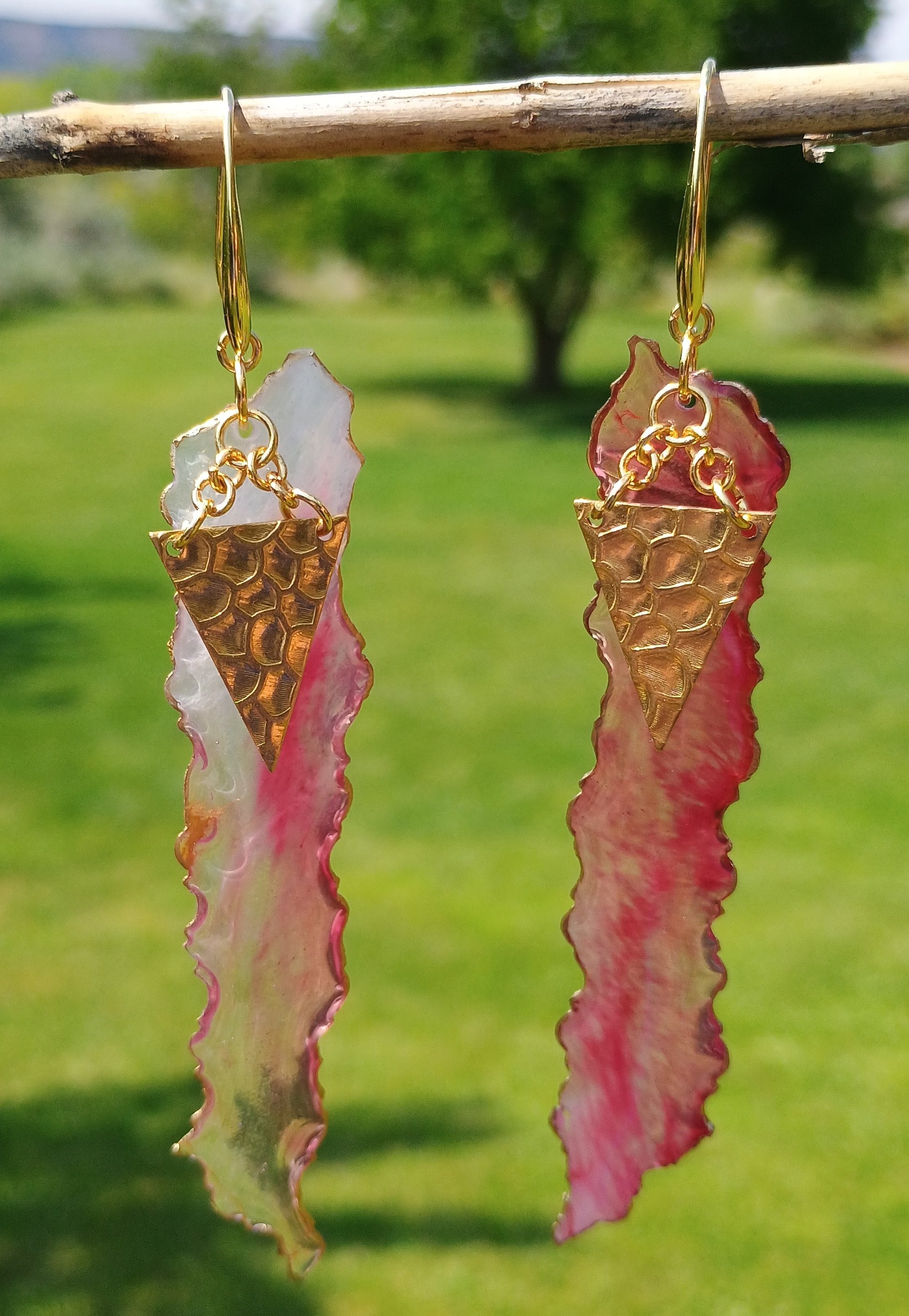 Gold and pink earrings hanging from a branch with a blurred green outdoor background