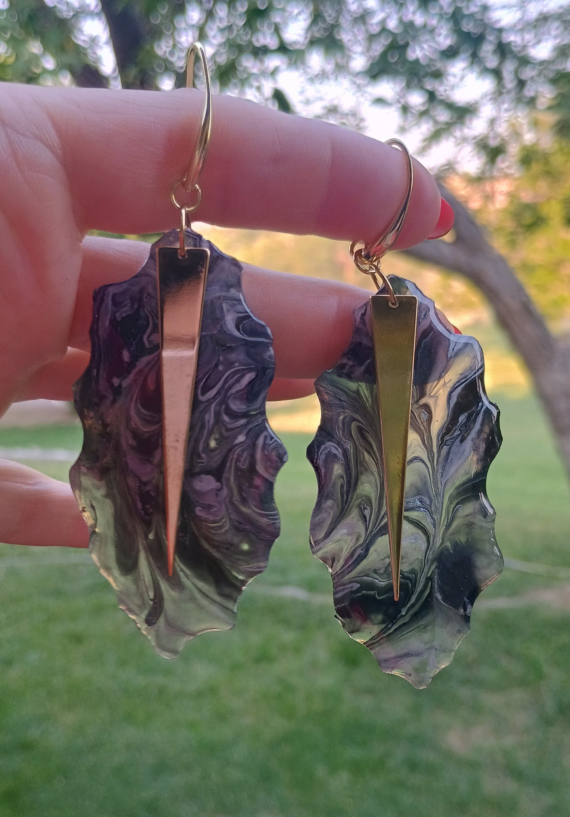 Hand-moulded black and gold abstract resin dangle earrings held between fingers outdoors, showing swirling black, grey, and white resin with an 18K gold triangle accent and gold ear wire hardware, one-of-a-kind handmade statement jewelry by Style By Jess at Gotta Have It Design