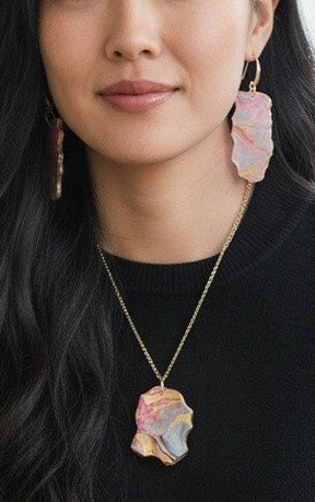 A woman with long dark hair sits in a wooden chair in a bright room, wearing a black long-sleeve top and black pants, accessorized with a gold necklace featuring a pastel multicolor flower pendant and matching drop earrings.