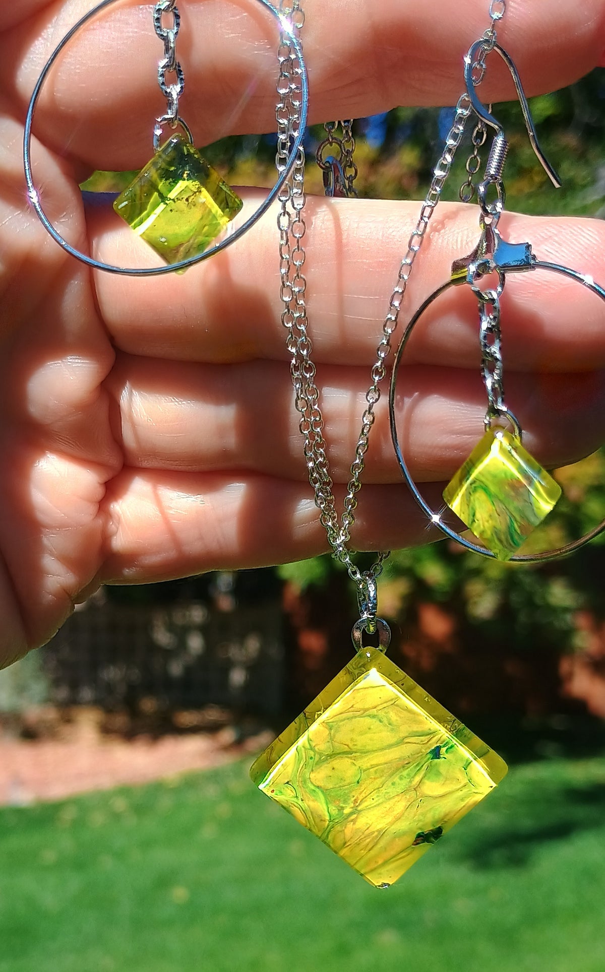 A hand holding up silver hoop earrings and a matching chain necklace with translucent yellow-green diamond-shaped pendants against an outdoor background of green grass and trees.